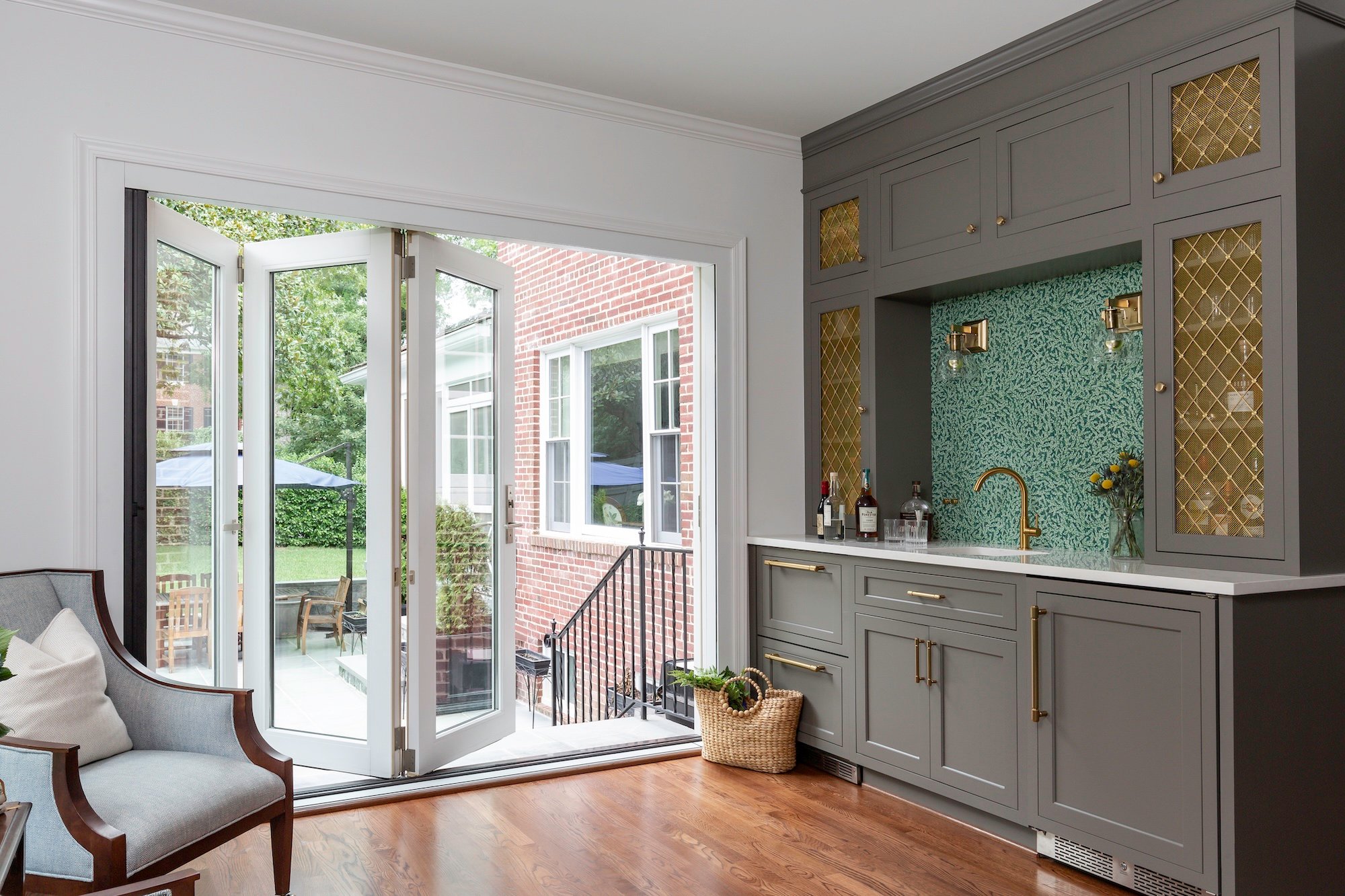 Award-winning remodeler project in Northwest DC featuring elegant gray built-in bar cabinetry with gold accents, glass-front cabinets, mosaic backsplash, and French doors opening to a brick patio and outdoor living space.