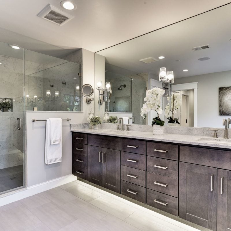 This modern bathroom, crafted by an award-winning remodeler in Bethesda, showcases a large mirror above a double-sink vanity with dark wood cabinets and silver handles. A glass shower enclosure sits to the left, while white orchids and towels add elegance. Recessed lighting completes the ambiance.