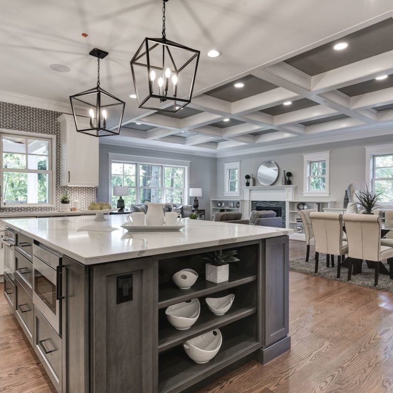 Modern kitchen in Northwest DC with a large island featuring built-in shelves and drawers, topped with a white countertop. Two geometric pendant lights hang above. The background shows a dining area with a table and chairs, and large windows providing Washingtons natural light.