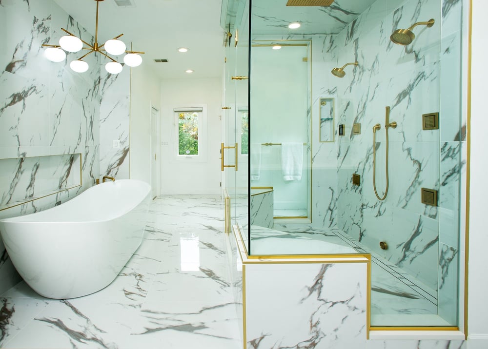 Experience award-winning design in this luxurious Potomac bathroom, featuring white and gray marble walls and floors, a sleek freestanding bathtub, modern gold-accented fittings, a glass-enclosed shower, and a contemporary chandelier.