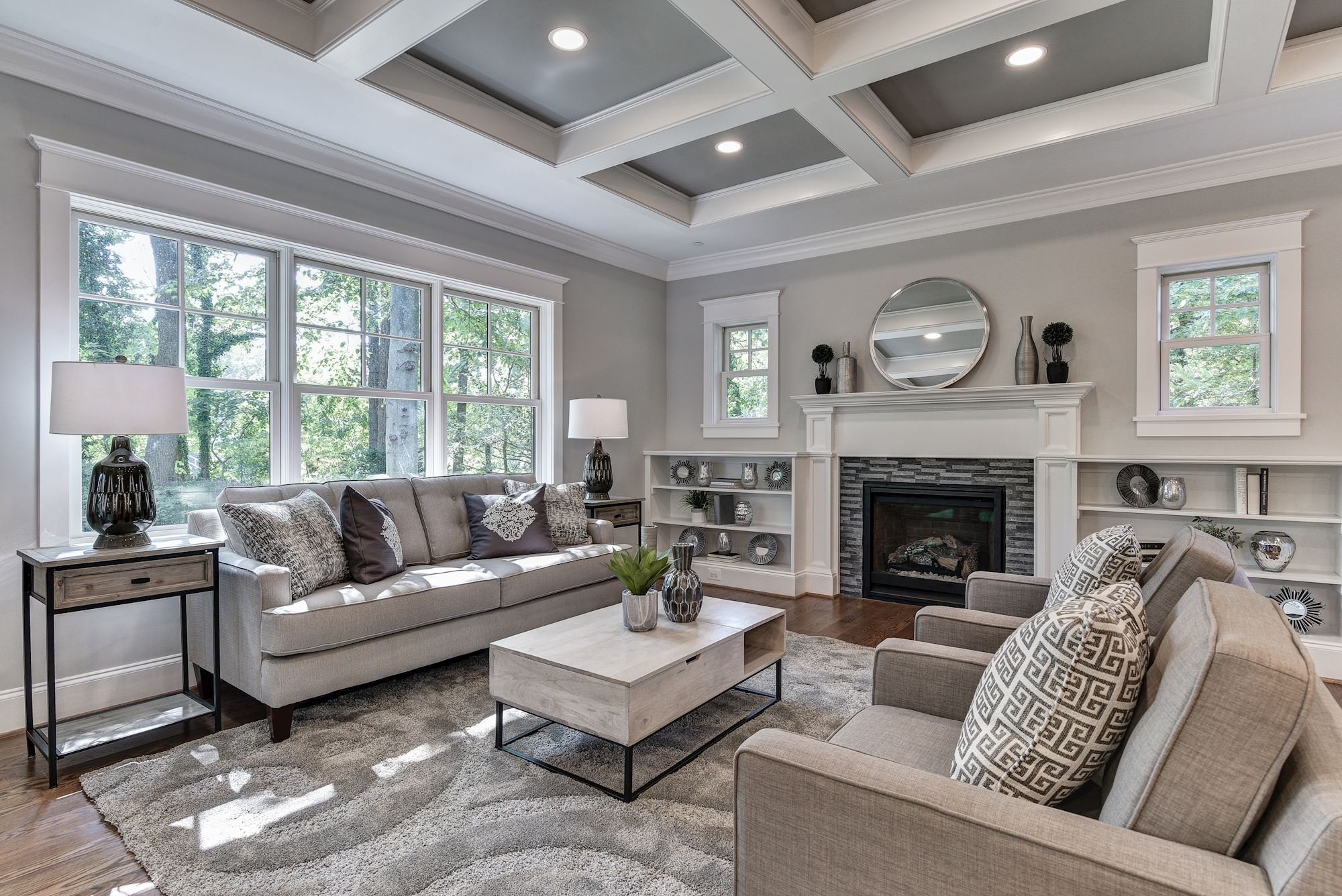 "An award-winning modern living room in Bethesda features gray sofas, a light patterned rug, a coffee table, a fireplace with a round mirror above it, built-in shelves, and large windows letting in natural light.