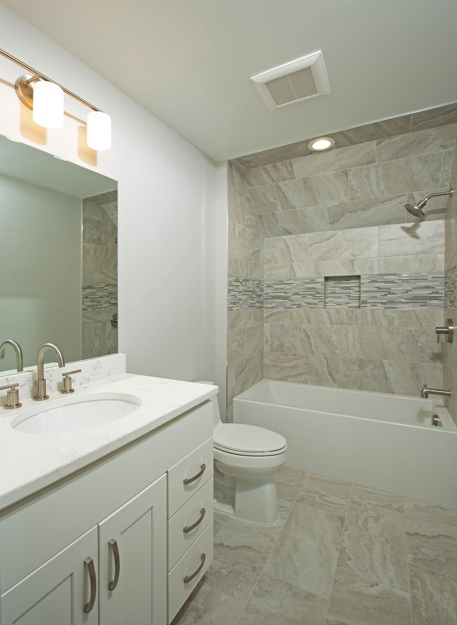 <img alt="Modern bathroom in Bethesda with white vanity, marble countertop, and silver fixtures. The award-winning remodeler designed the bathtub and shower area with beige stone tiles, a decorative accent strip, large mirror, and overhead light above the sink." src="#" />