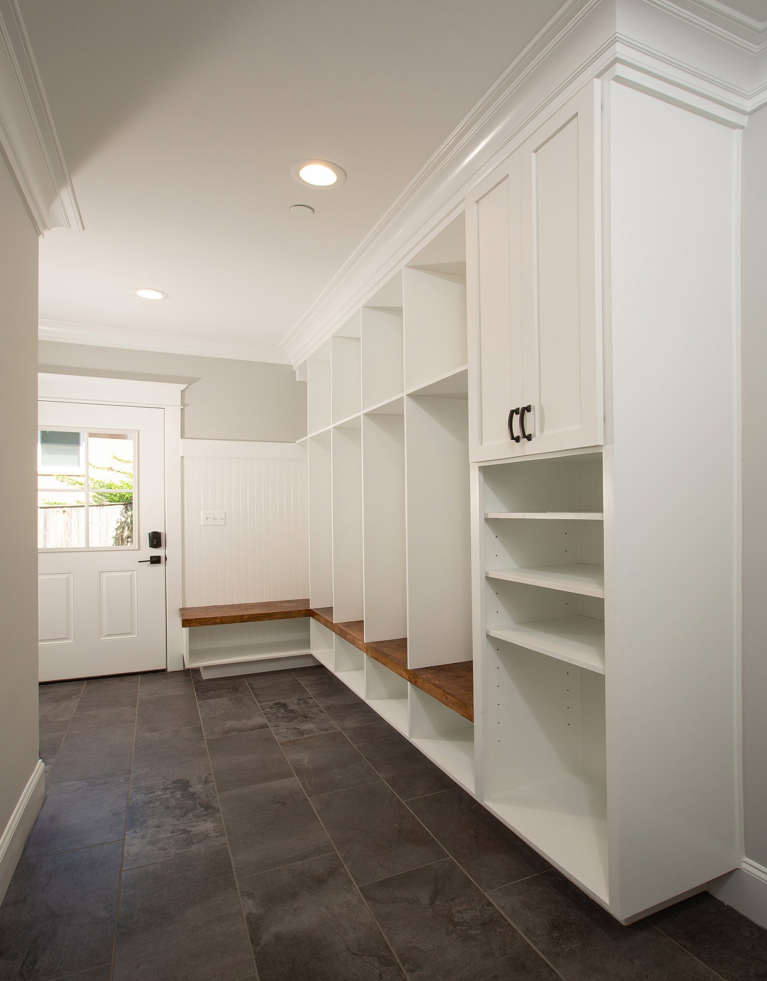 <img alt="A modern mudroom in Bethesda with white built-in storage cabinets, open shelves, and wooden bench seating. The floor is tiled in dark gray, and a white door with glass panels accents this award-winning remodelers design." src="#" />