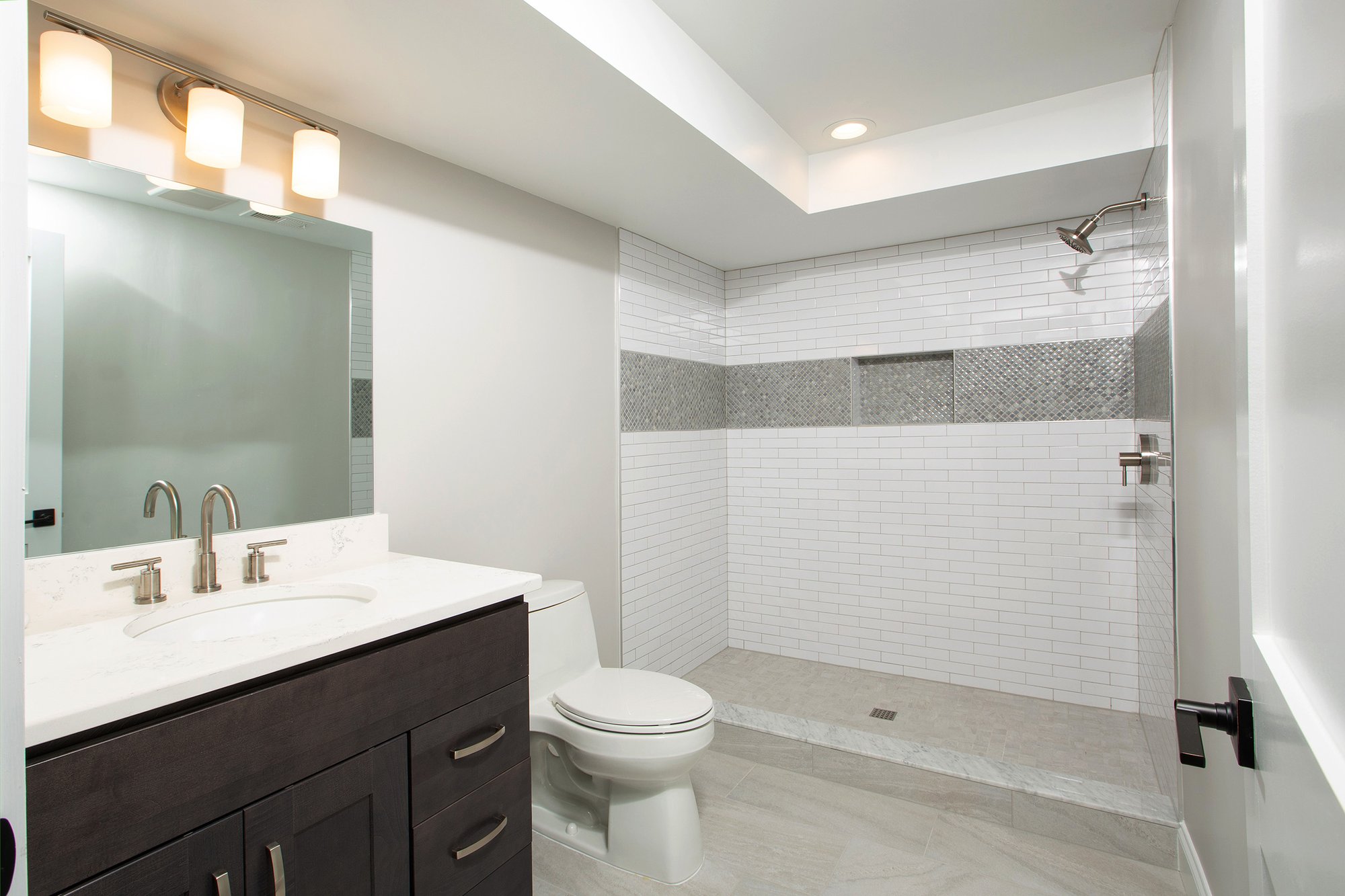 <img alt="Modern bathroom in Bethesda with white vanity, marble countertop, and silver fixtures. The award-winning remodeler designed the bathtub and shower area with beige stone tiles, a decorative accent strip, large mirror, and overhead light above the sink." src="#" />