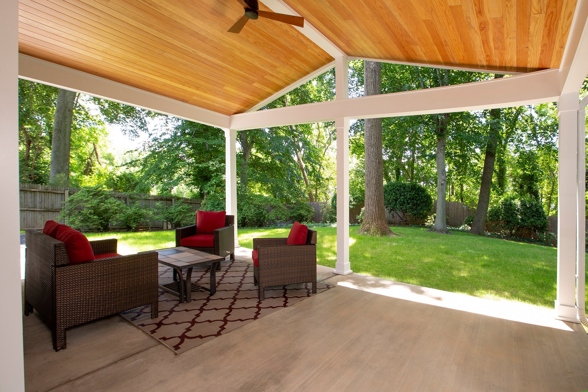 <img alt="Award-winning covered patio in Bethesda featuring a wooden ceiling, outdoor wicker furniture with red cushions, a patterned rug, and views of a lush green backyard with trees and sunlight." 