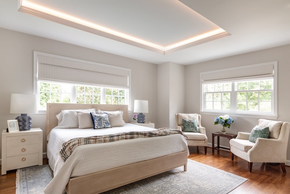 Award-winning remodel in Northwest DC showcasing a serene primary bedroom with tray ceiling lighting, upholstered bed, soft neutral tones, and a cozy seating area by the windows.