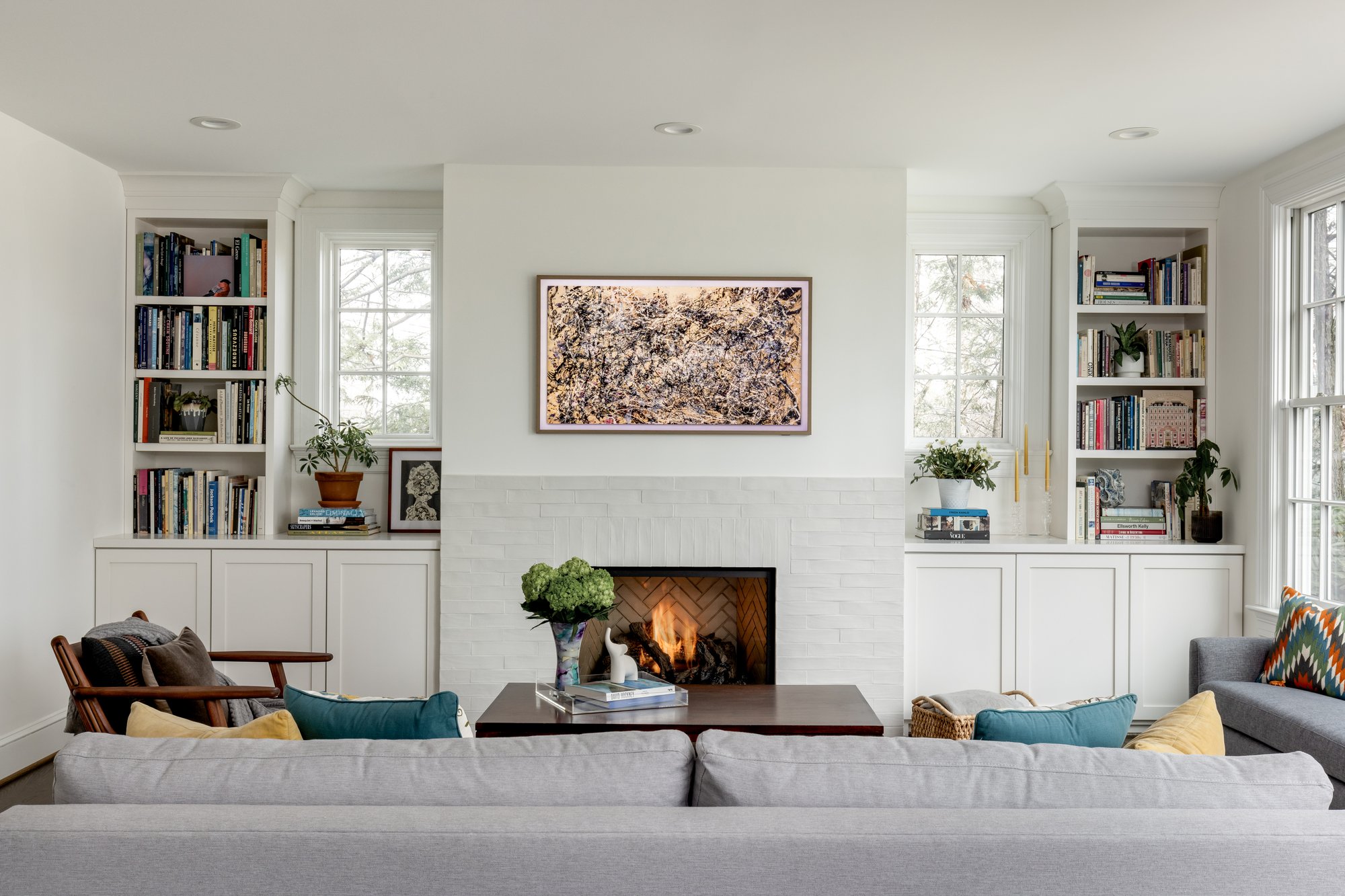 <img alt="A bright, modern living room by award-winning remodeler Smiley Renovations in Northwest DC features a gray sofa, armchairs, a coffee table, built-in shelves with books, a fireplace, and large windows. Above the fireplace is framed earthy-toned art." src="#" />