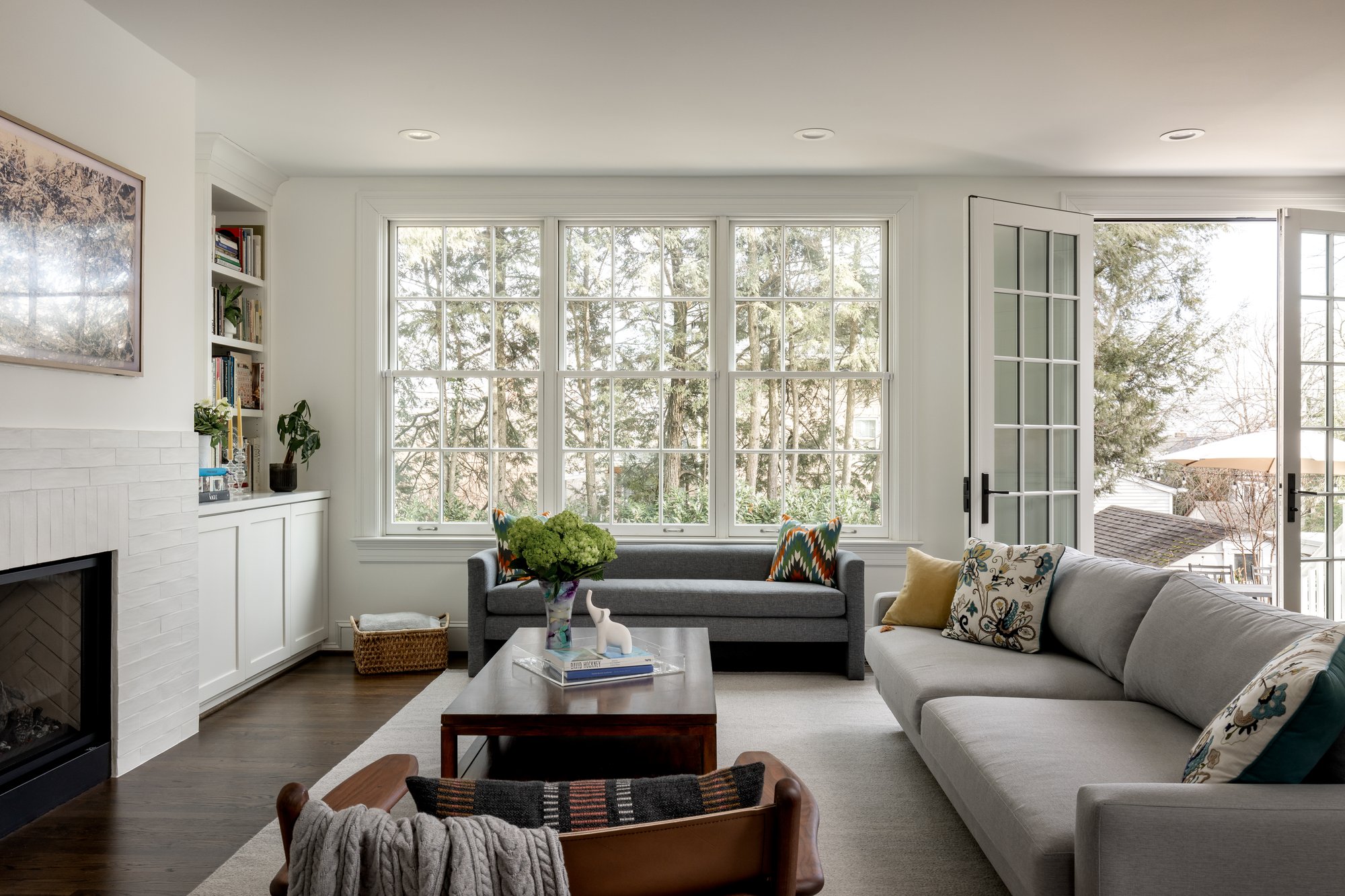 <img alt="A bright living room in Northwest DC by Smiley Renovations features large windows, a gray sofa, a dark gray loveseat, a wooden coffee table with books and flowers, a fireplace, built-in shelves, and double doors opening to the outside." src="#" />