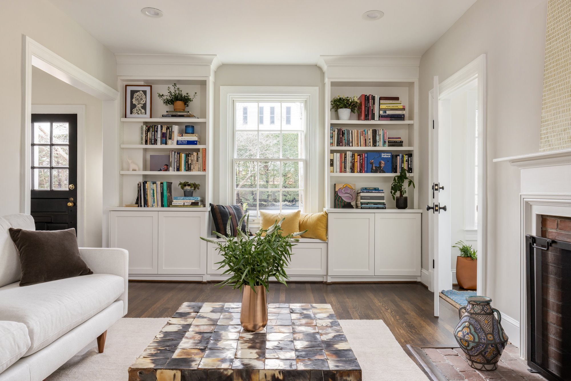 <img alt="Bright living room with built-in white bookshelves, a window seat with pillows, and a fireplace—expertly crafted by Smiley Renovations, an award-winning remodeler serving Northwest DC." src="#" />