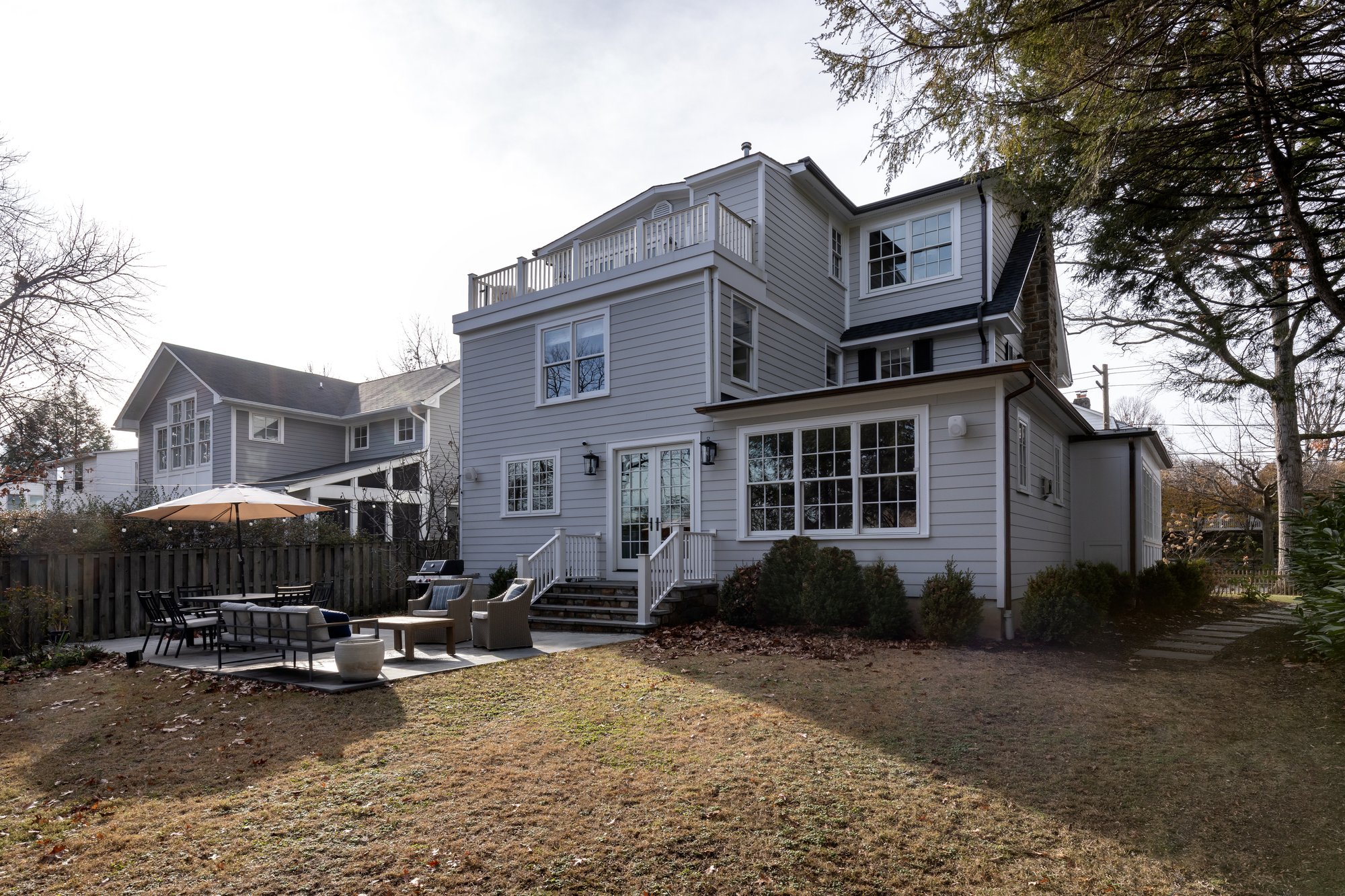<img alt="A two-story gray house with white trim, a stone chimney, large windows, and a small fenced yard with shrubs—beautifully updated by award-winning remodeler Smiley Renovations—situated on a residential street in Northwest DC under a partly cloudy sky." src="#" />