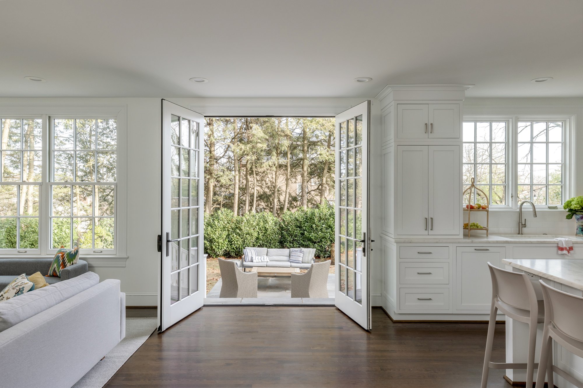 <img alt="An award-winning, bright modern kitchen with white cabinets and a breakfast bar opens through double french doors to a patio with outdoor seating, surrounded by greenery and trees. Large windows bring in natural light." src="#" />