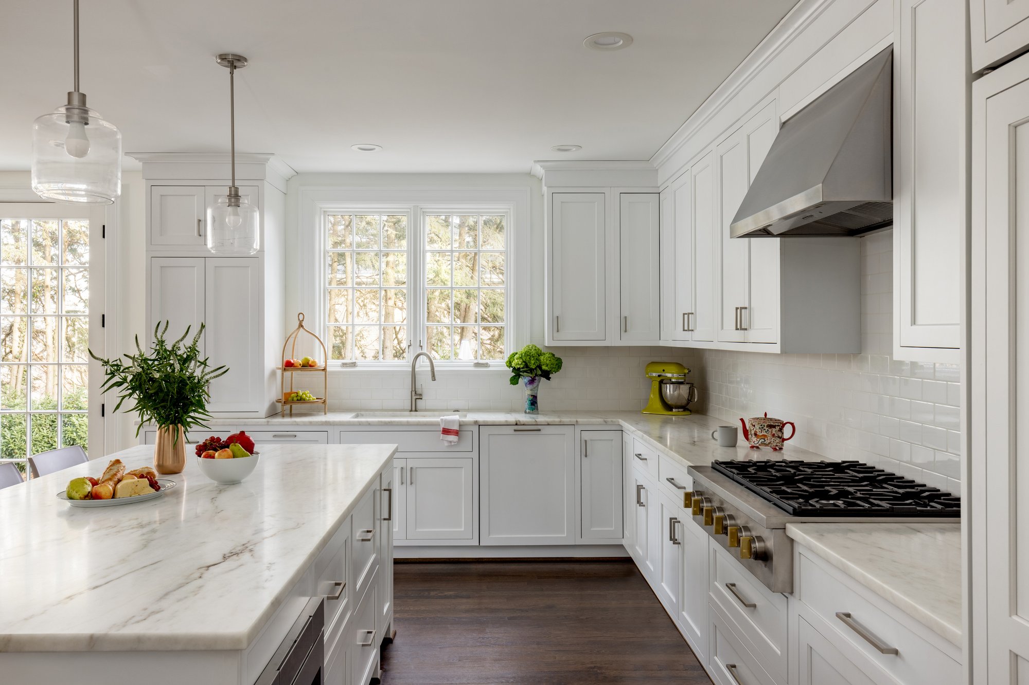 <img alt="Modern white kitchen by Smiley Renovations featuring an island, four white chairs, pendant lights, built-in appliances, and hardwood floors. Countertops display fruit bowls and plants. Natural light pours in from a window on the left." src="#" />