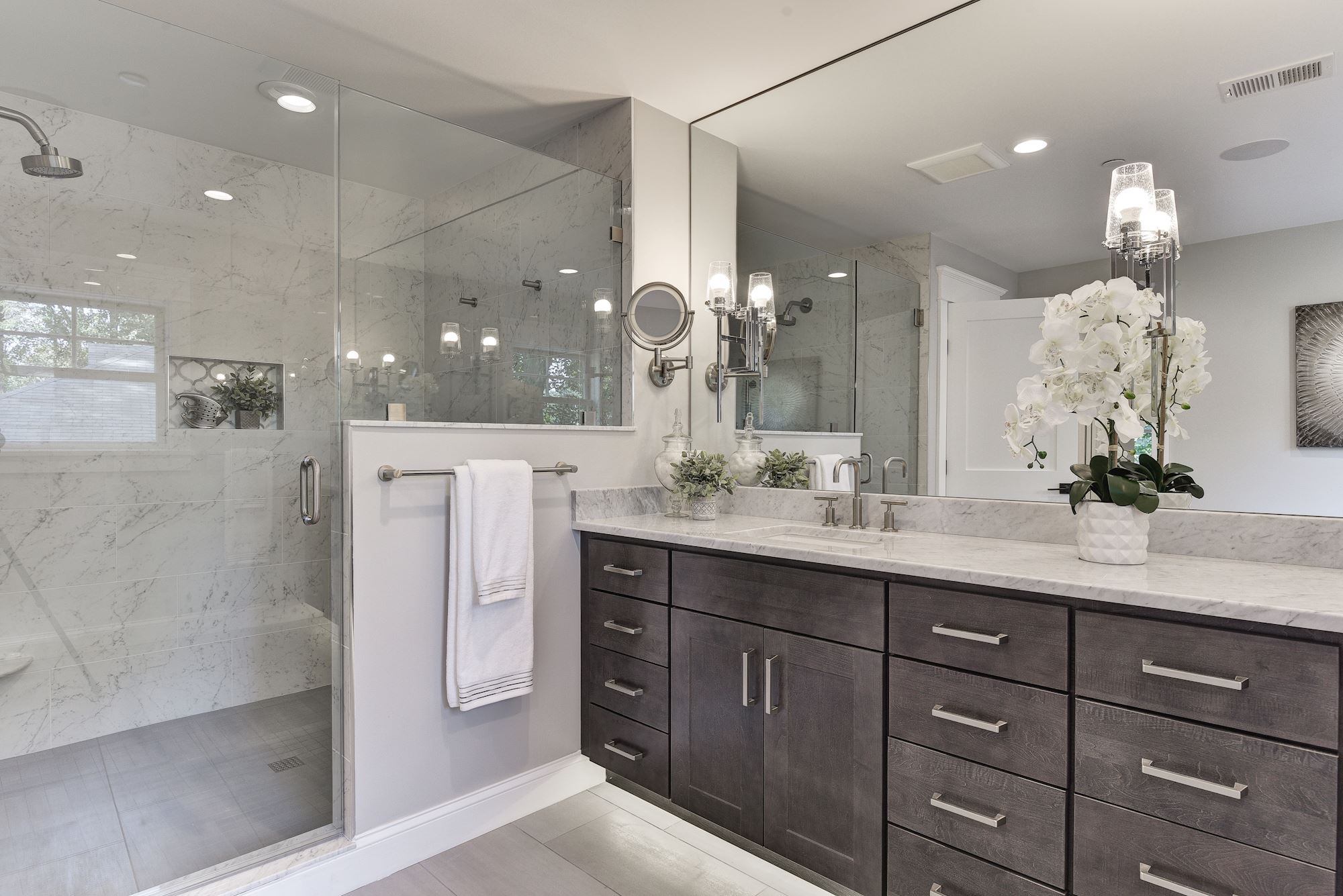 A modern bathroom, part of a recent home remodel, boasts a large glass-enclosed shower on the left with marble walls. On the right, a dark wood vanity with a marble countertop holds twin sinks and a potted orchid. A large mirror reflects the room's elegant details.