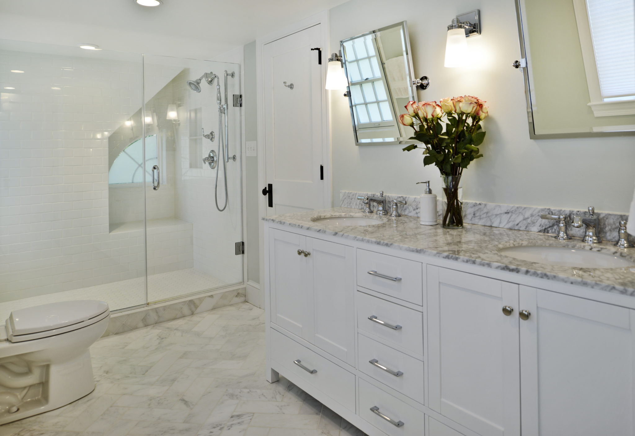 A bathroom featuring a shower and sink, with tiled walls and floor. The space includes a mirror above the sink, cabinetry for storage, and a vase as decor.