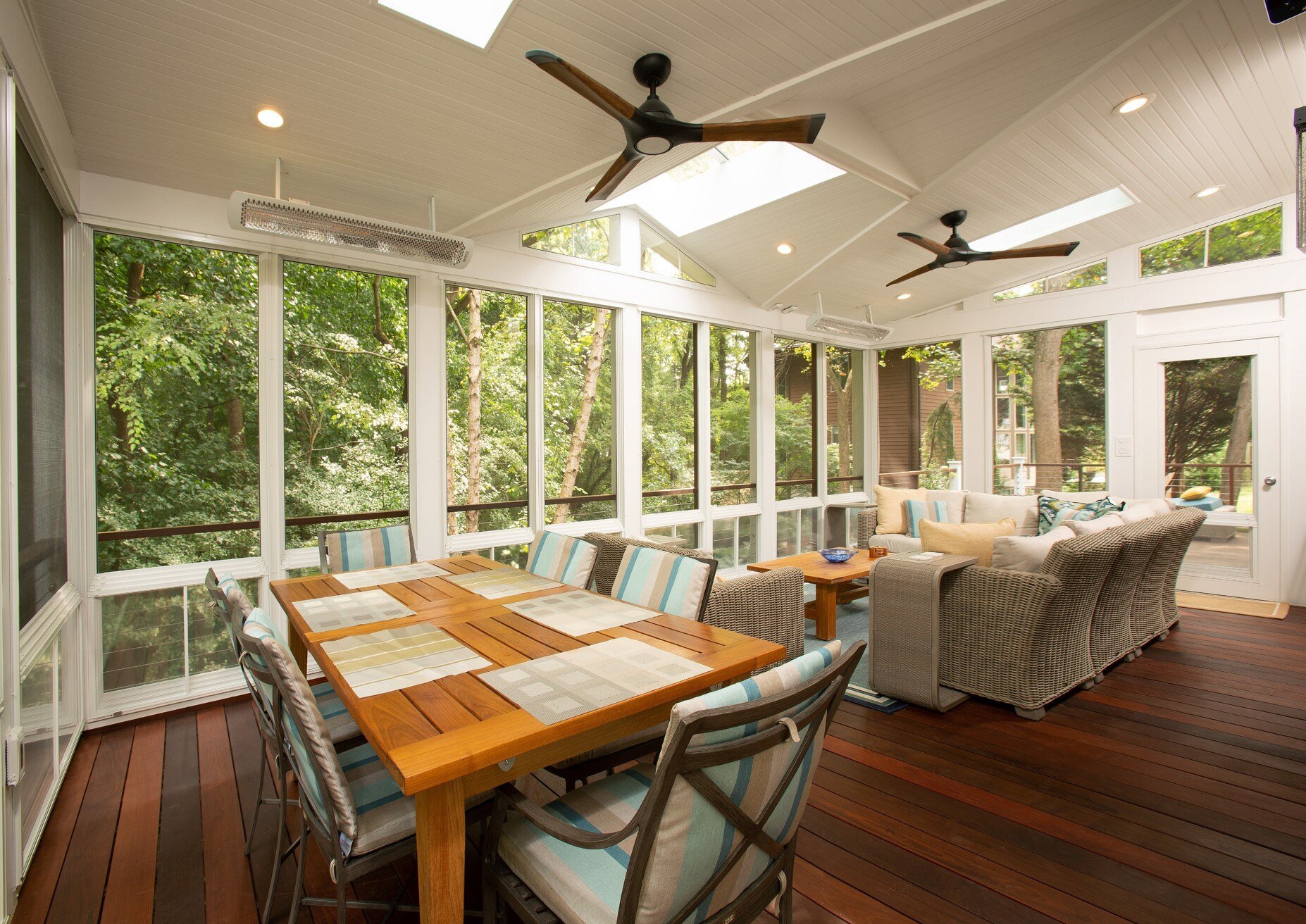 A bright, screened-in porch with wood flooring showcases a Montgomery County remodel, featuring a large dining table with placemats, cushioned chairs, ceiling fans, and sofas. Large windows reveal green trees outside.