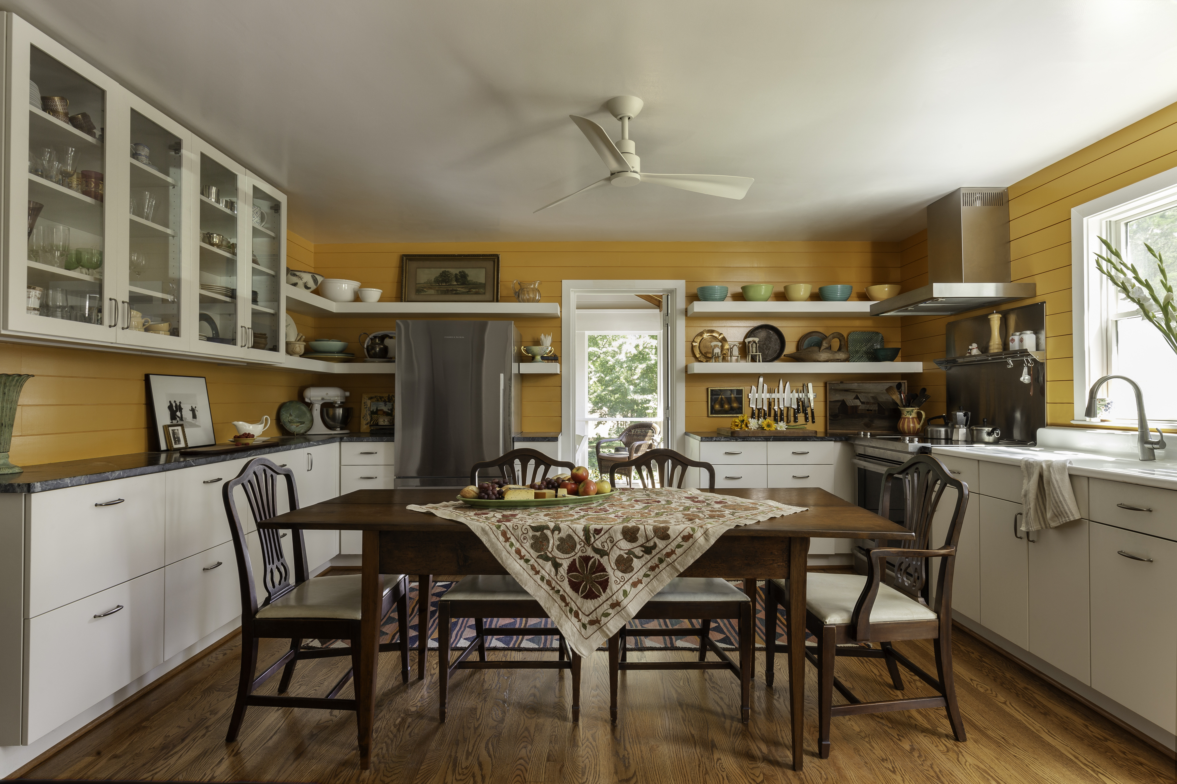 A bright kitchen featuring a dining table surrounded by chairs, with cabinetry and countertops visible. The room includes a sink, cupboards, and appliances, with a window allowing natural light to illuminate the space. The floor is designed for indoor use, complementing the overall interior design of the home.