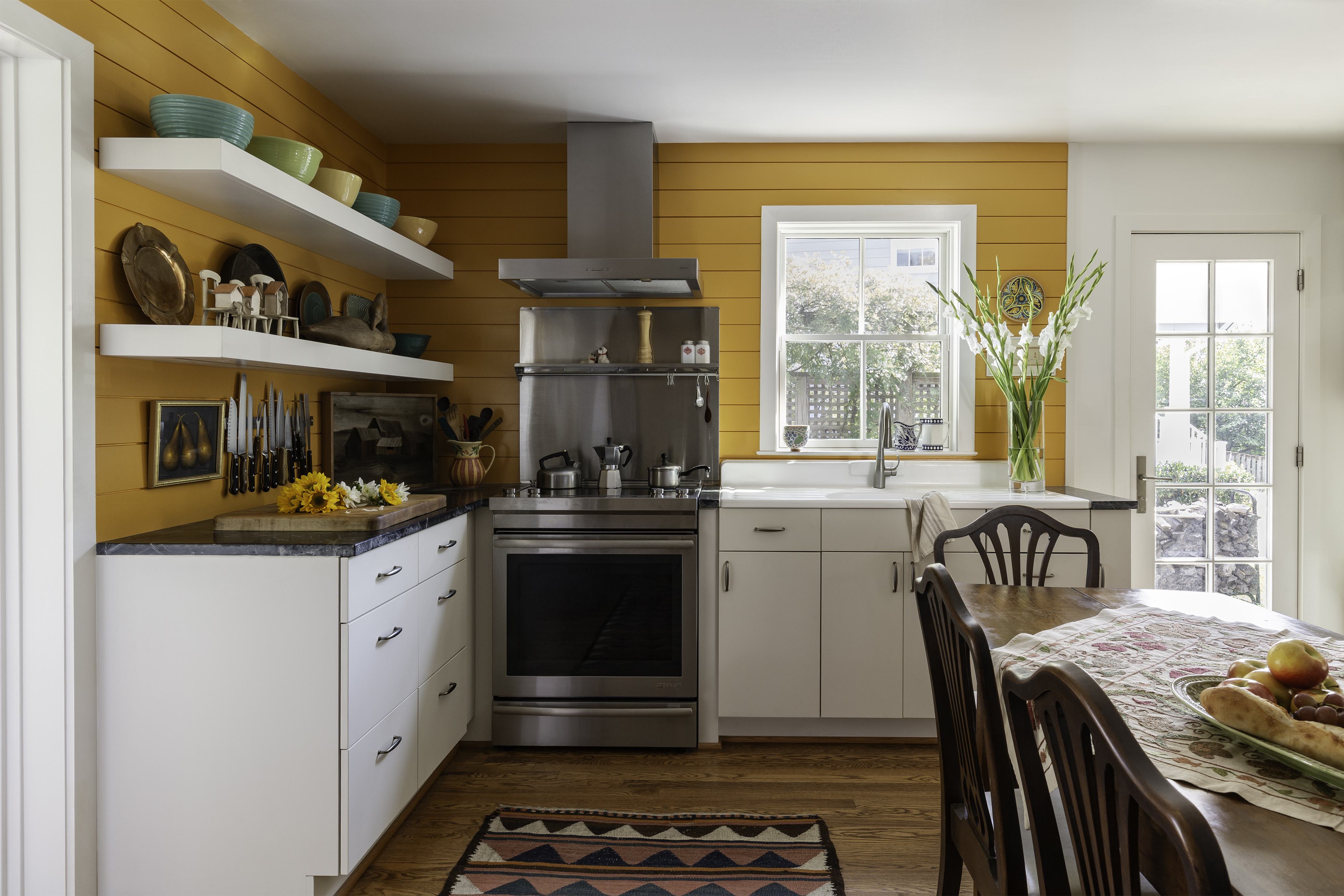 A kitchen featuring a stove, oven, sink, and cabinetry. The space includes countertop surfaces, a refrigerator, and an exhaust hood above the stove. There are windows allowing natural light in and furnishings like chairs. The overall interior design reflects a cozy home environment.