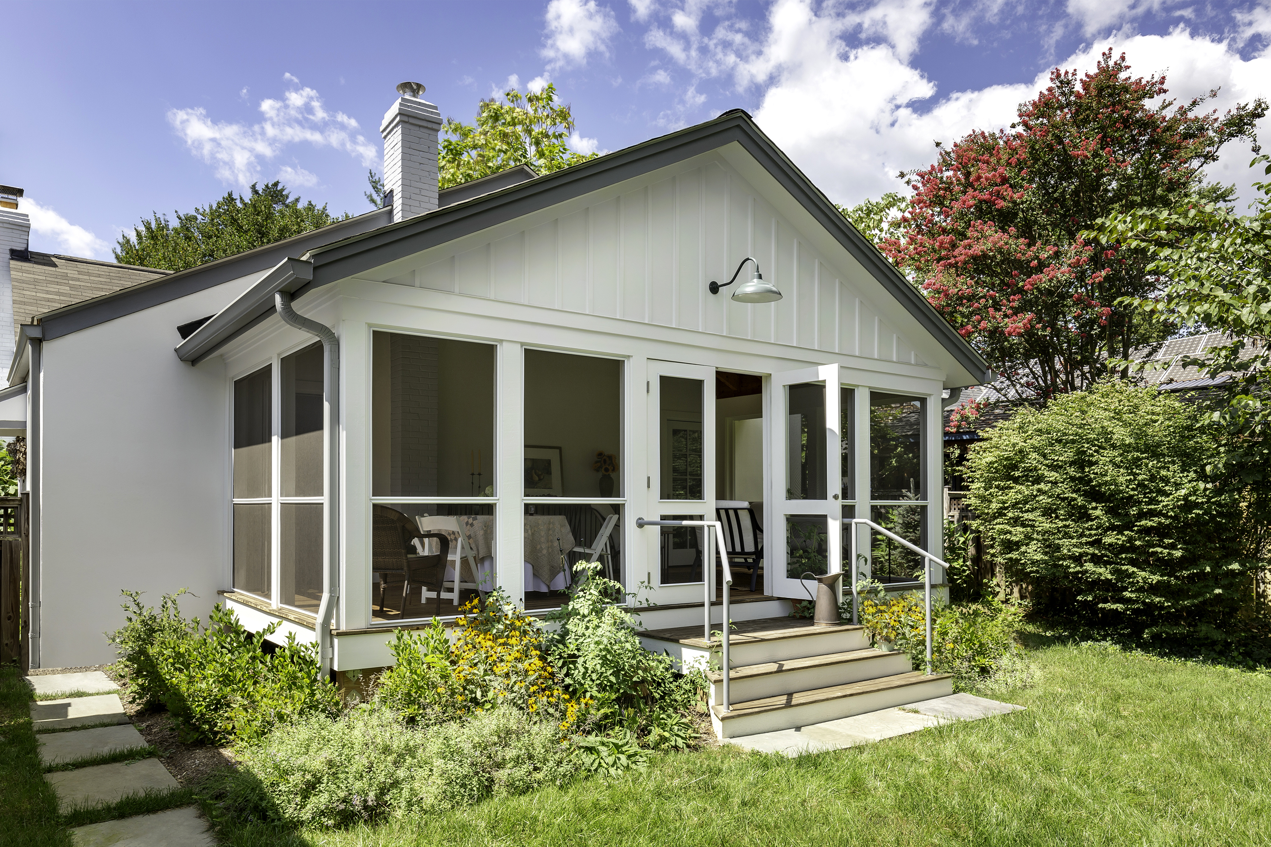 A cozy outdoor room featuring a wooden table surrounded by chairs, with hardwood floors and a ceiling fan overhead. The walls are adorned with molding, and there's a window allowing natural light to fill the space. The design reflects an inviting atmosphere typical of an attic or loft setting.