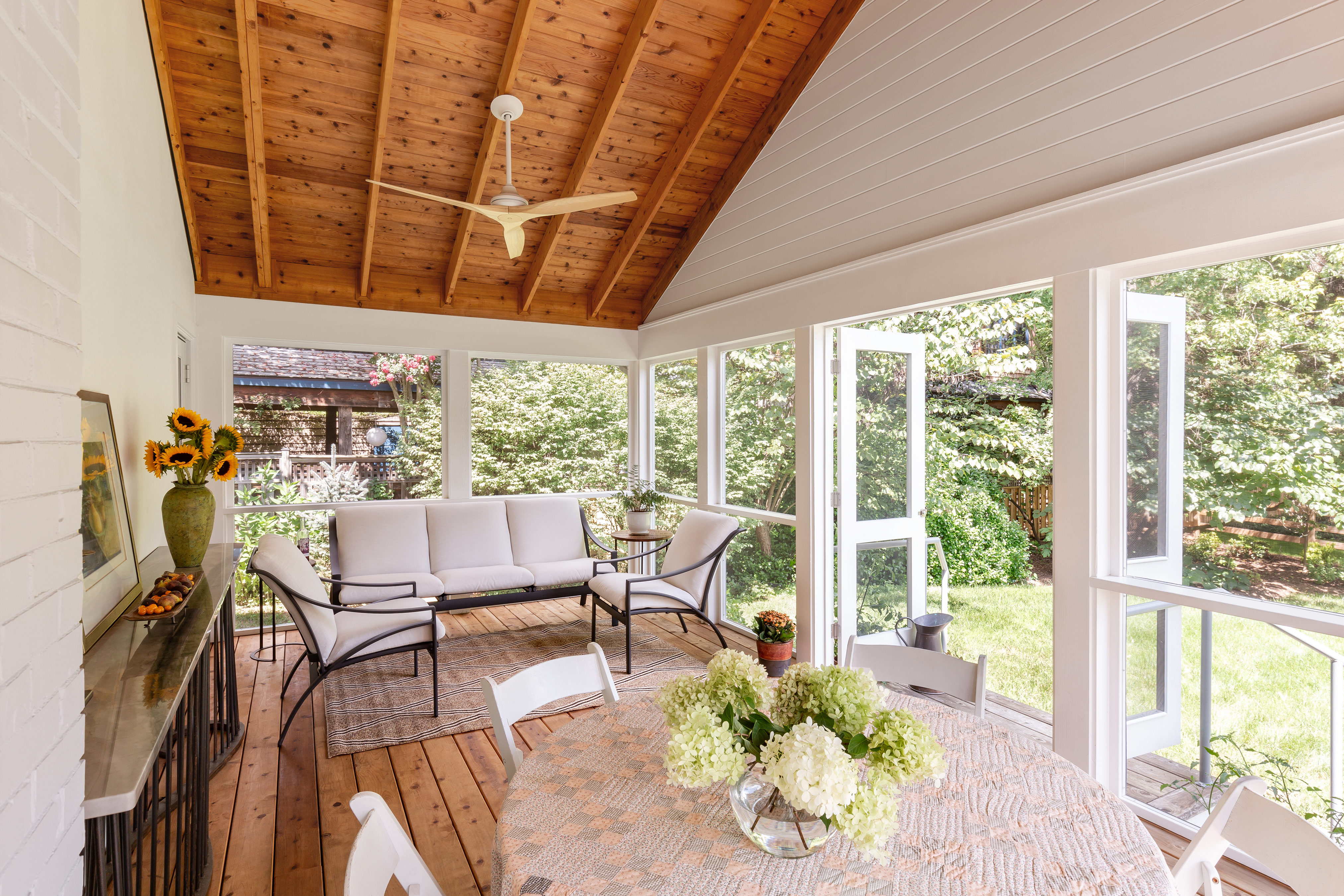 A cozy outdoor room featuring a wooden table surrounded by chairs, with hardwood floors and a ceiling fan overhead. The walls are adorned with molding, and there's a window allowing natural light to fill the space. The design reflects an inviting atmosphere typical of an attic or loft setting.