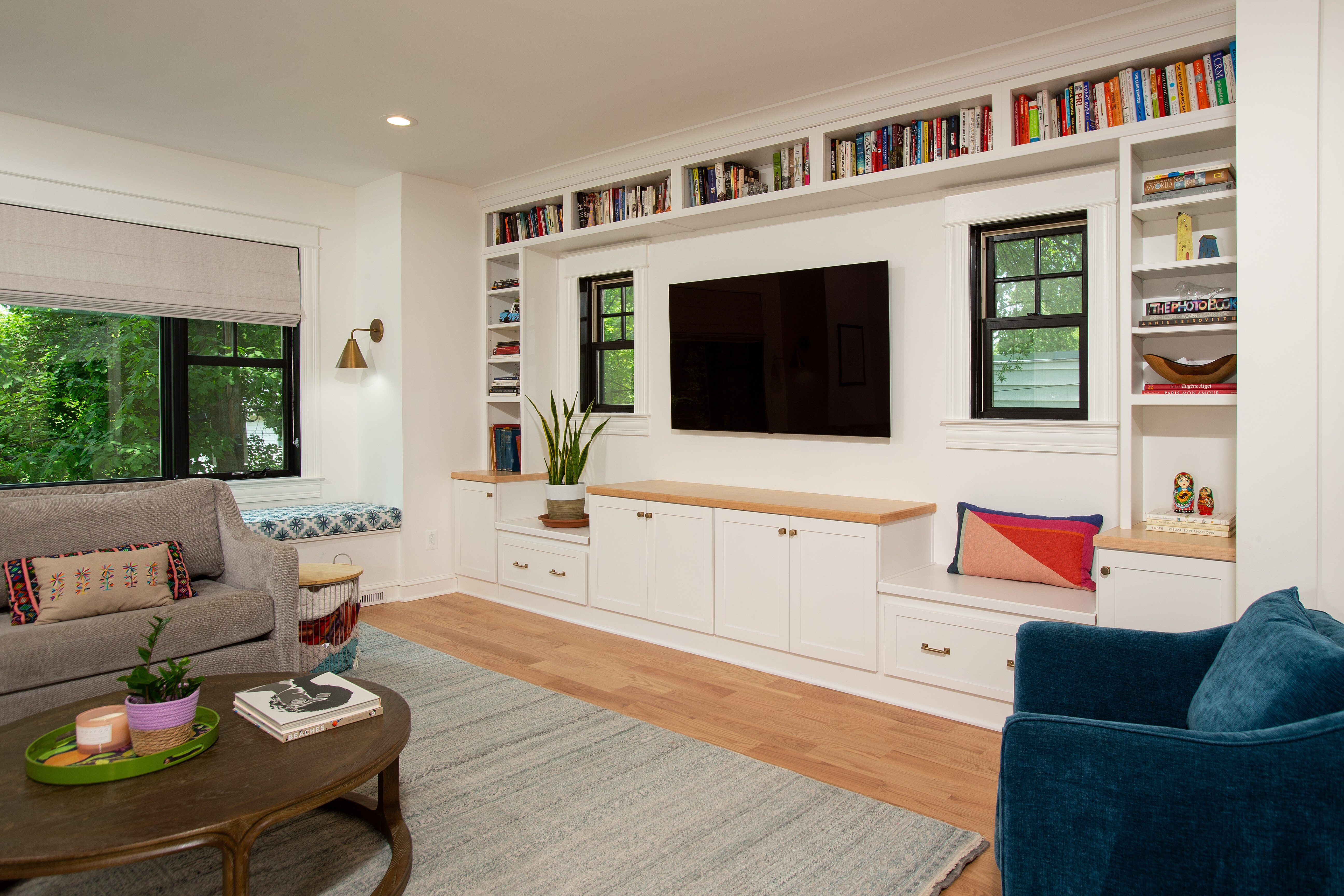 A cozy living room, part of an award-winning remodel in Montgomery County, features a large TV on a white built-in shelf surrounded by books. The space includes a gray sofa, blue armchair, round wooden coffee table, and beige rug on a wooden floor. Natural light pours through two windows.