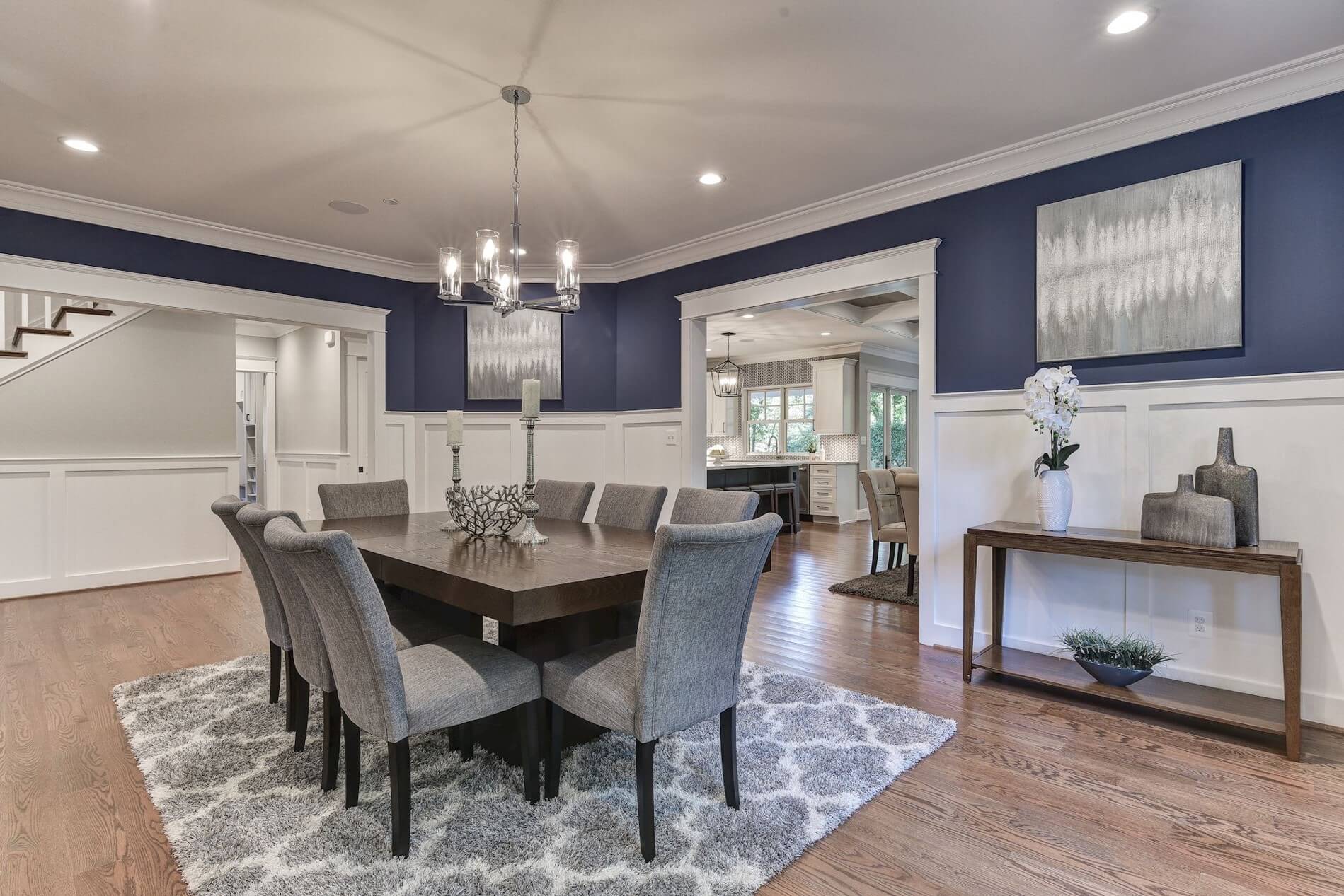 Dining room designed by a Bethesda remodeling expert features a dark wooden table with eight gray upholstered chairs on a patterned rug. Dark blue walls and white wainscoting complement the chandelier, while art and decor adorn the walls and console table. Stairs and another room are visible.