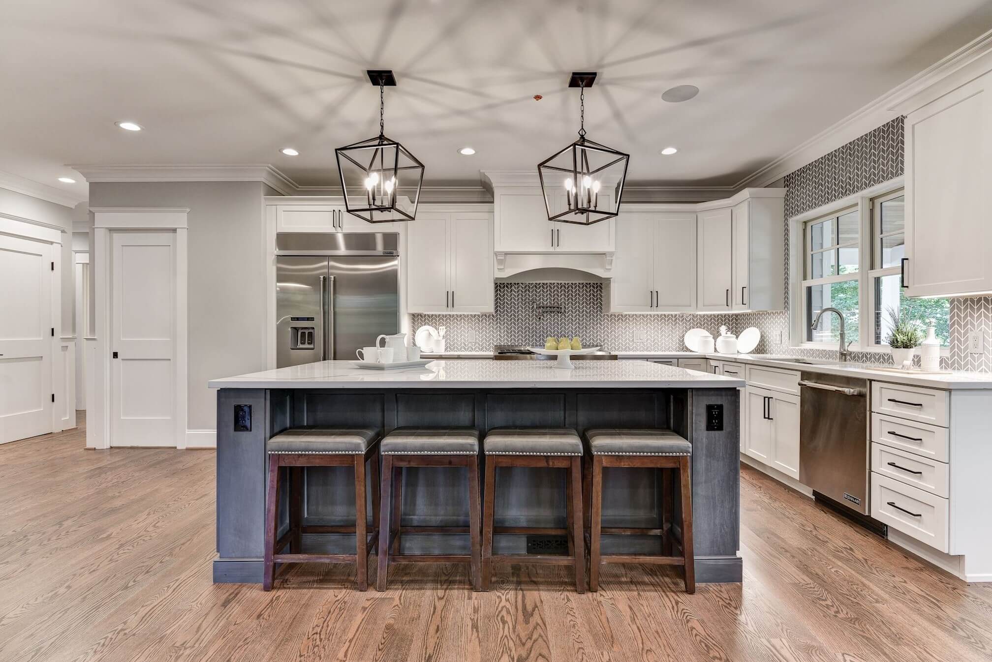 Modern kitchen in an award-winning remodeler project features white cabinets, a large island with a marble countertop, and four wooden stools. Geometric pendant lights hang above the island. Stainless steel appliances and a patterned backsplash complete this Montgomery County space with hardwood floors throughout.