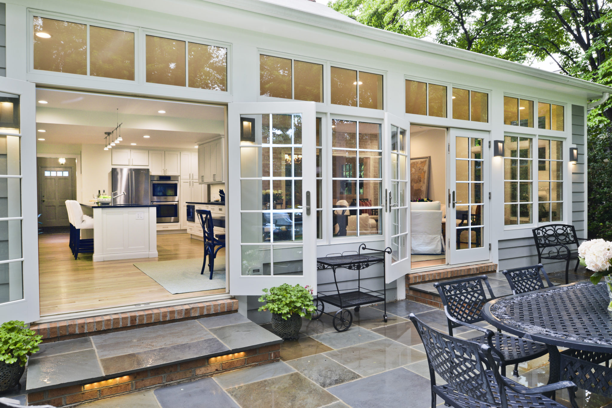 A bright kitchen and dining area with white cabinets and an island is visible through open glass doors, showcasing Montgomery Countys award-winning remodeler. The patio features a metal table and chairs, stone flooring, and potted plants, with trees in the background.