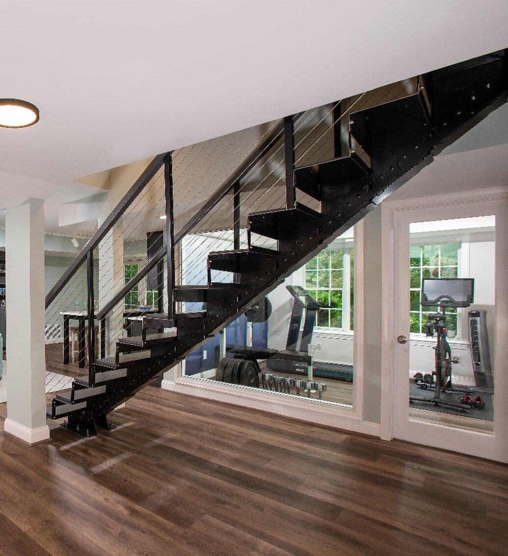 A modern interior showcases an award-winning remodel in Montgomery County, featuring an open, black staircase with metal railings leading to an upper floor. Below the stairs, large windows reveal a home gym with various exercise equipment on dark wood laminate flooring.