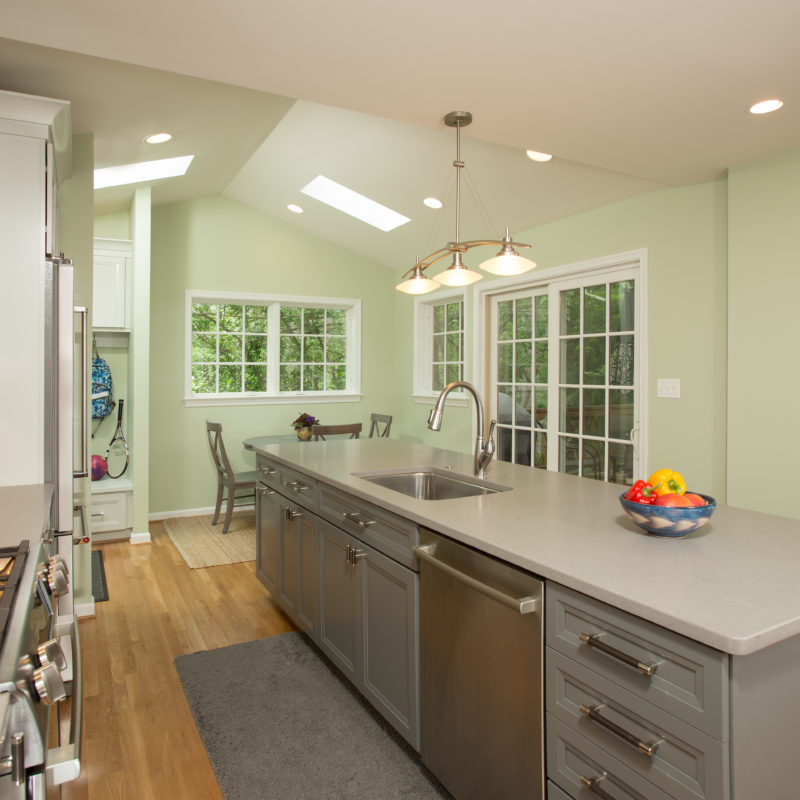 A bright kitchen featuring white cabinets, a countertop, sink, large skylights and window. The room has wood flooring and modern plumbing fixtures, creating a clean and inviting space.