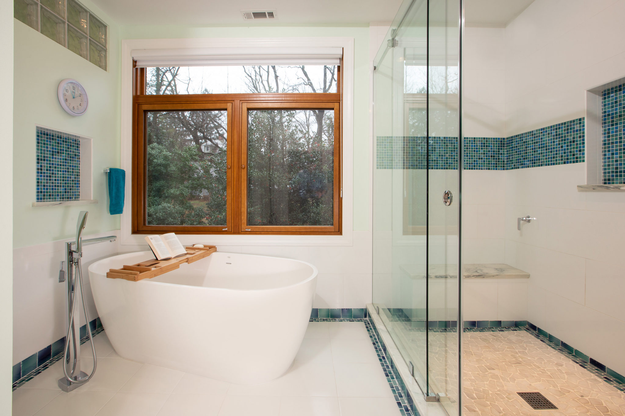 A bathroom featuring a free-standing bathtub and a sink, with tiled walls, a mirror, and various plumbing fixtures. The room includes a window, a large shower with a glass door and a bench.