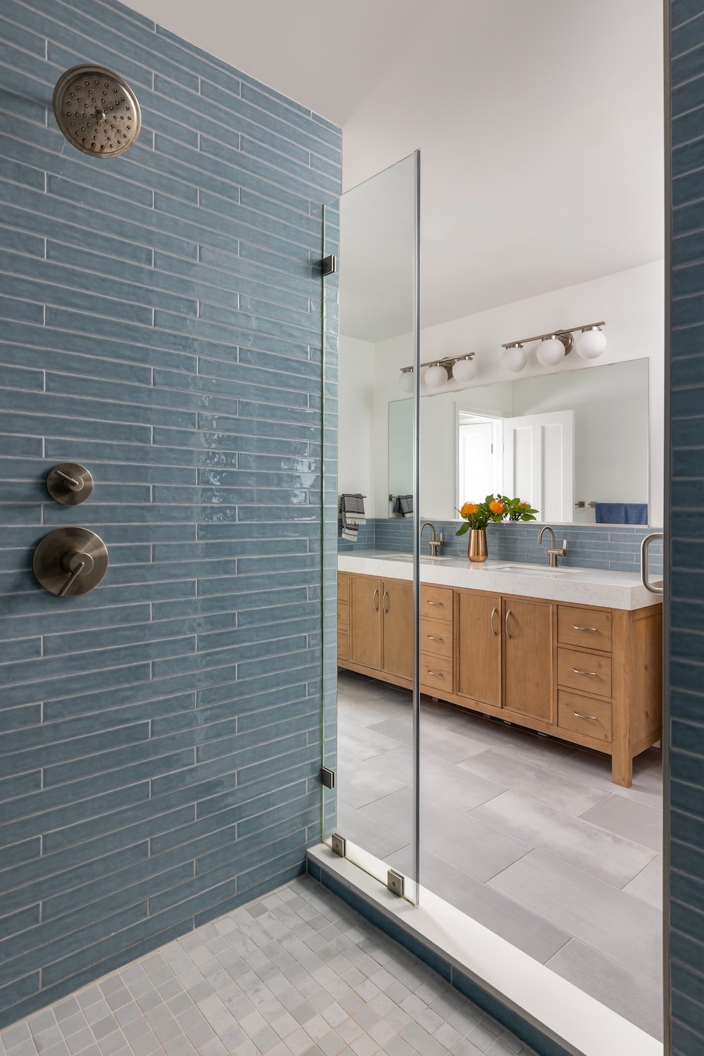 Modern bathroom in Northwest DC with blue subway tile shower, glass door, double-sink wooden vanity, large mirror, and orange flowers. Designed by our award-winning team, white walls and silver fixtures complete the clean look.