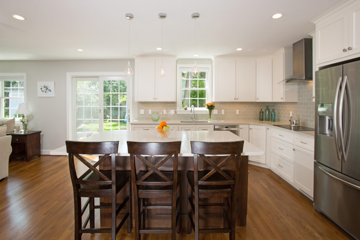 This award-winning remodeler has crafted a modern kitchen with white cabinets, stainless steel appliances, and a center island adorned by four dark wooden chairs. Pendant lights illuminate the space, while a window and glass doors provide natural light. A vase of flowers and a bowl of oranges complete this Montgomery County gem.