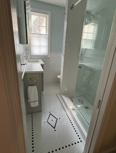 A narrow bathroom designed by an award-winning remodeler features a glass shower on the right, a small white vanity with a sink on the left, and a window with blinds above the toilet at the far end. The white mosaic floor with black diamond patterns enhances this stunning Montgomery County remodel.