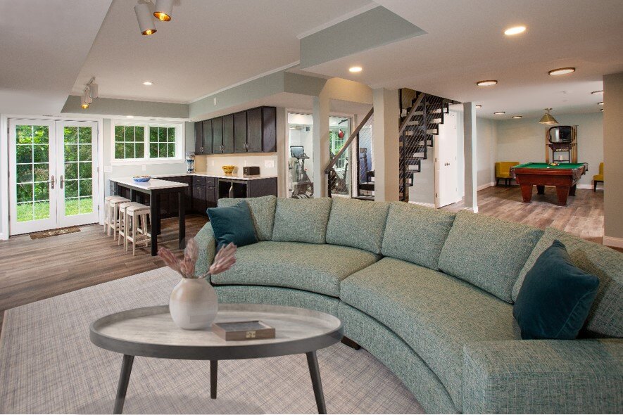 A modern open-concept living space in Chevy Chase features a light blue curved sofa, a round coffee table, and a decorative vase. In the background, a kitchen with dark cabinets complements the pool table and large glass doors leading outside.