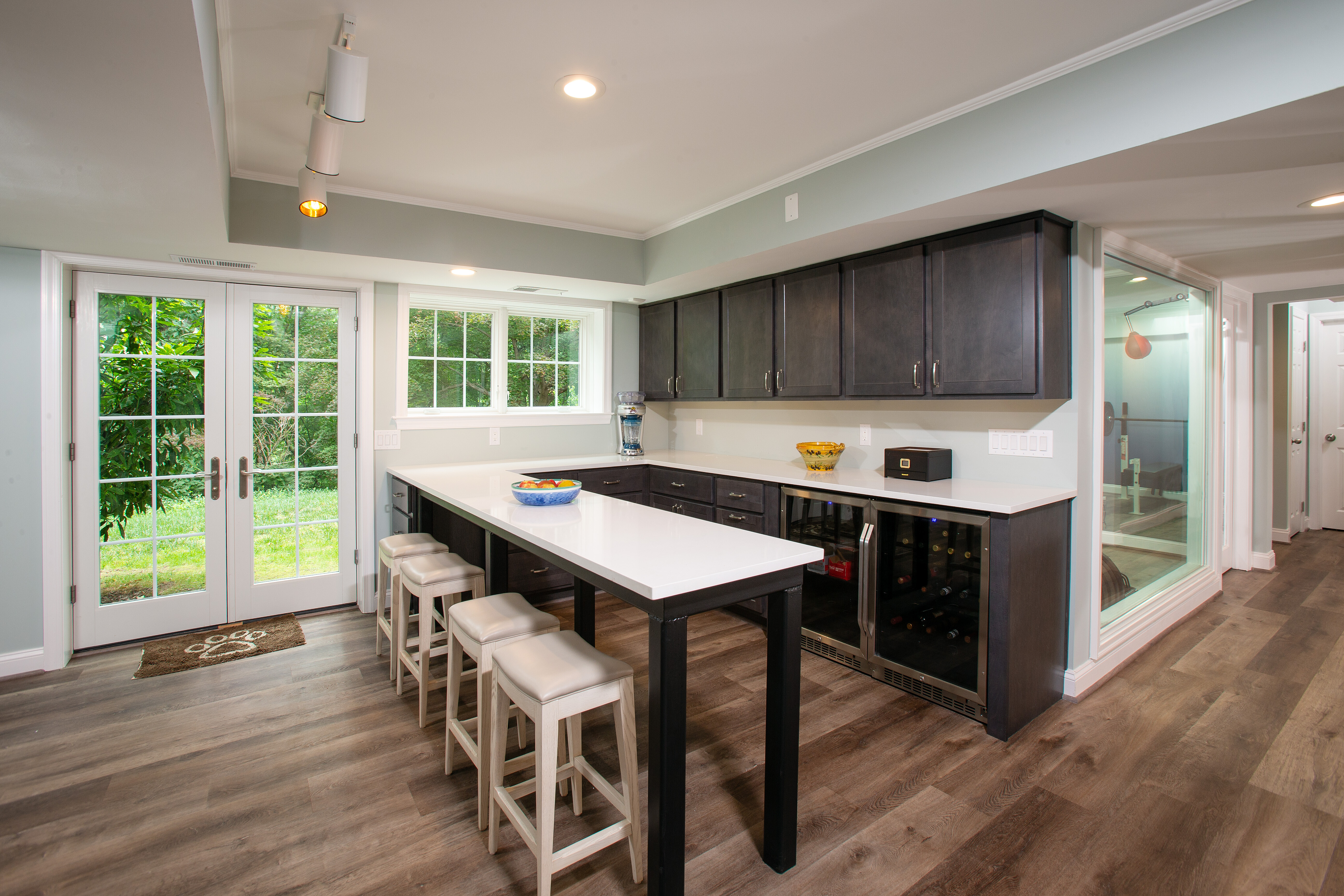 Spacious modern kitchen for Montgomery County homeowners, boasting a long island with white countertops and four matching stools. Dark cabinets contrast beautifully with light walls and floors. Large windows and French doors open to a lush outdoor area, while a glass wall reveals an adjacent room.