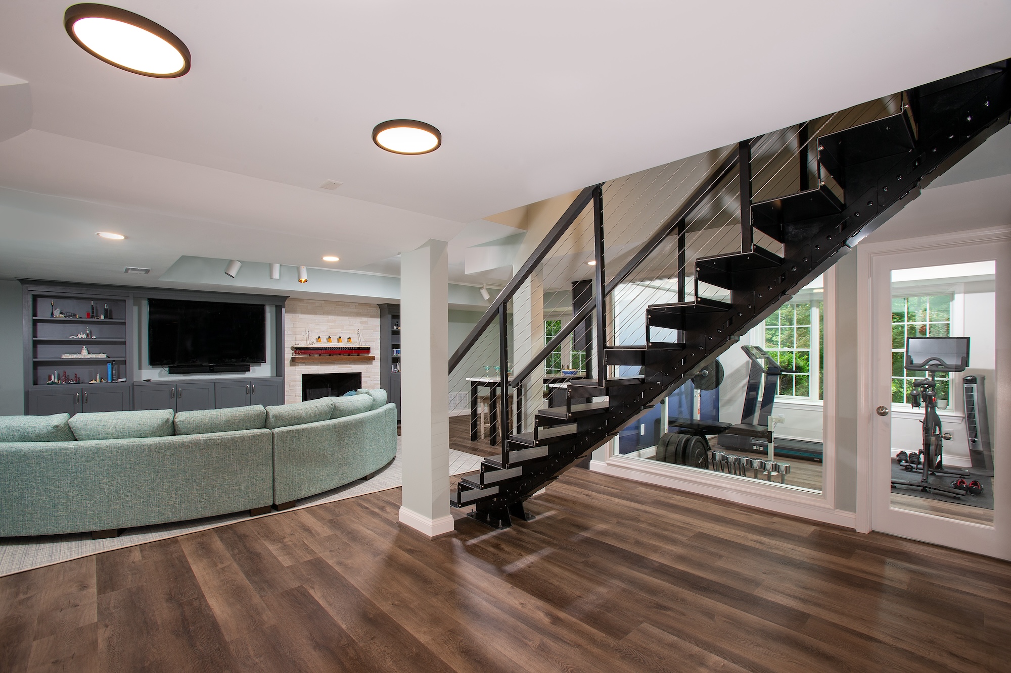 Modern Bethesda basement with wood flooring, a curved sofa facing the TV and fireplace, black open-riser staircase with glass railing, and a home gym visible through glass doors—crafted by an award-winning remodeler.