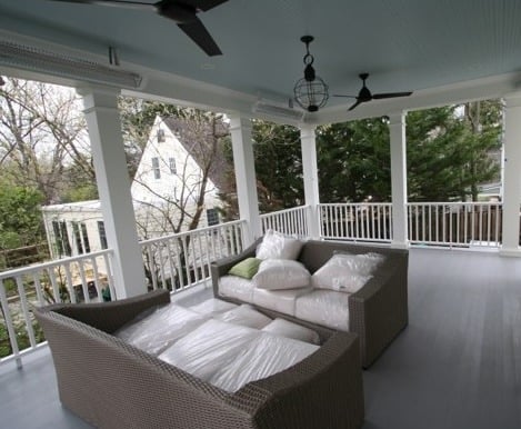 A covered porch crafted by an award-winning remodeler features a light blue ceiling and white pillars, showcasing a wicker sofa draped in plastic. Ceiling fans and decorative lighting grace the space, with trees and a charming Montgomery County house visible in the background.