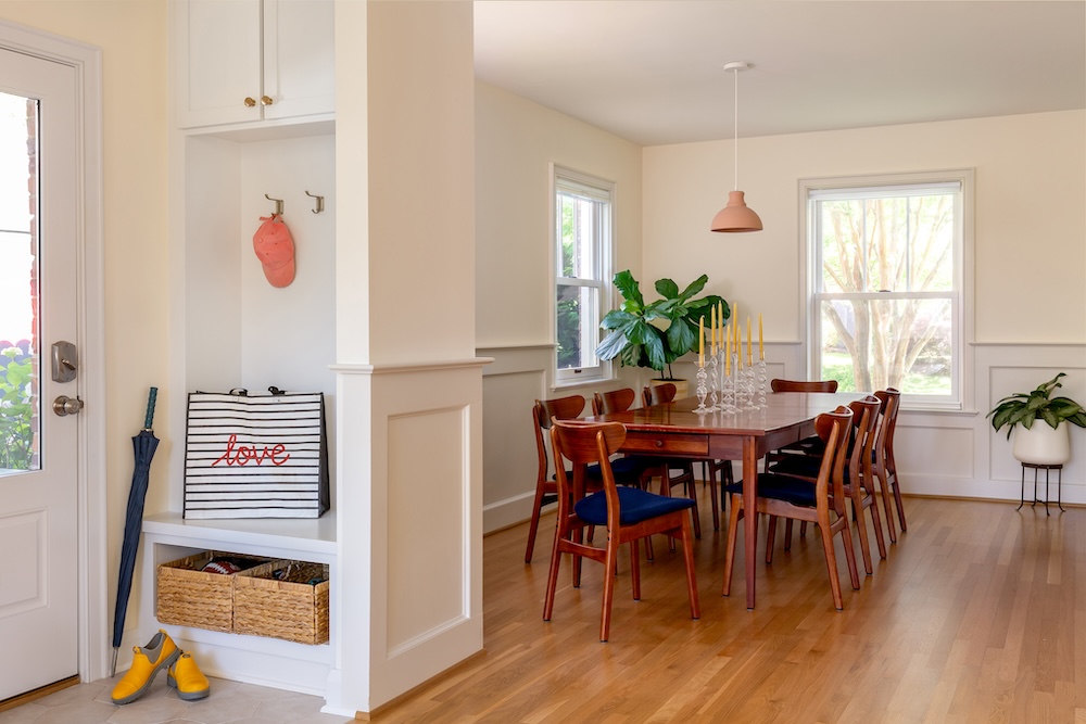 A bright dining room with a wooden table and eight chairs, large windows, potted plants, and a small entry area with an umbrella, boots, striped bag, and hanging hat. Sunlight fills the space. A bright dining room with a wooden table and eight chairs, large windows, potted plants, and a small entry area with an umbrella, boots, striped bag, and hanging hat. Sunlight fills the space.
