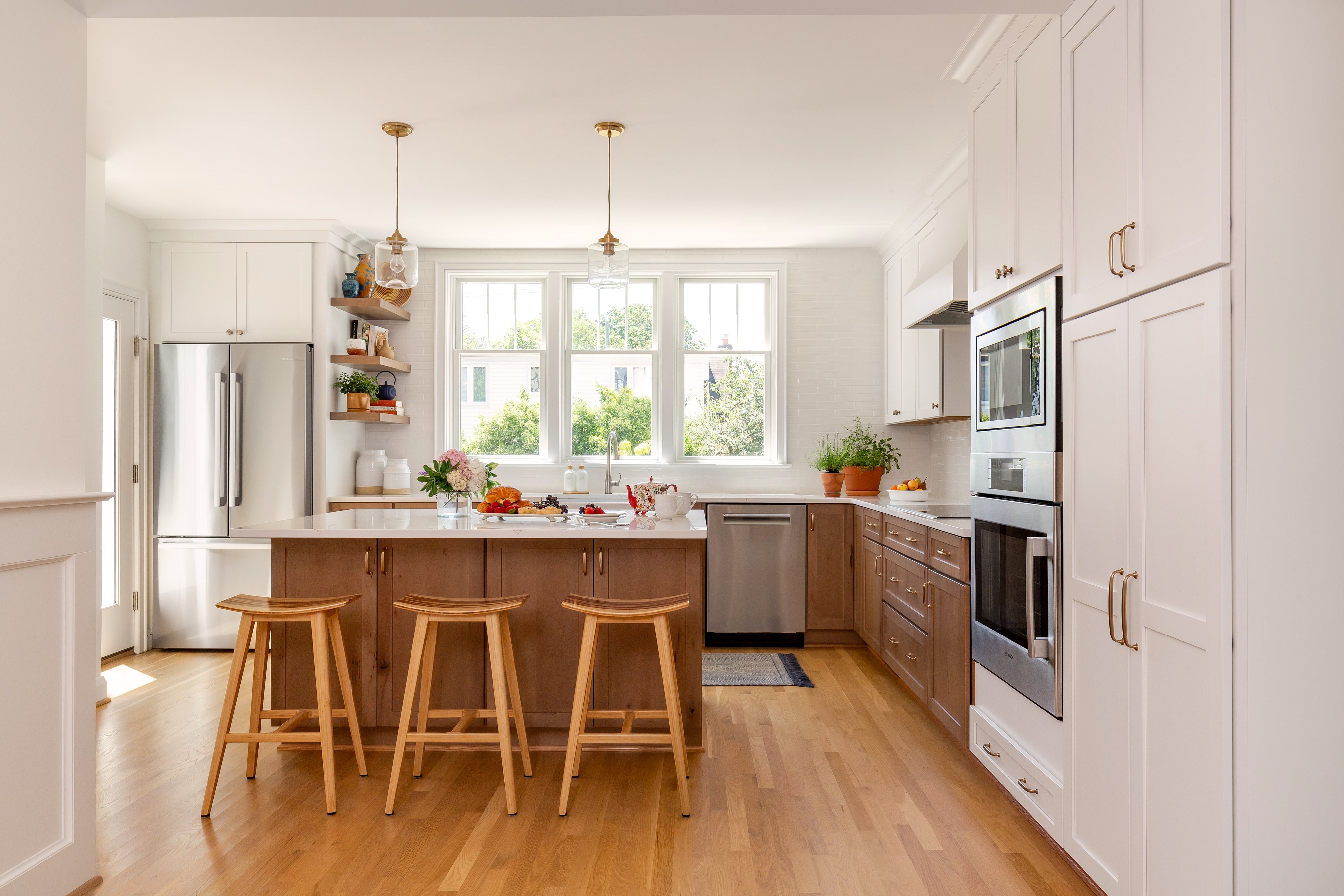 A modern kitchen in Montgomery County features wooden floors, a central island with three stools, pendant lights, and white cabinets. Appliances include double ovens and a refrigerator. Large windows provide natural light, while potted plants add greenery from an award-winning remodeler.