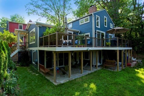 A two-story blue house with wooden deck, surrounded by trees and grass, featuring large windows and a red building nearby.