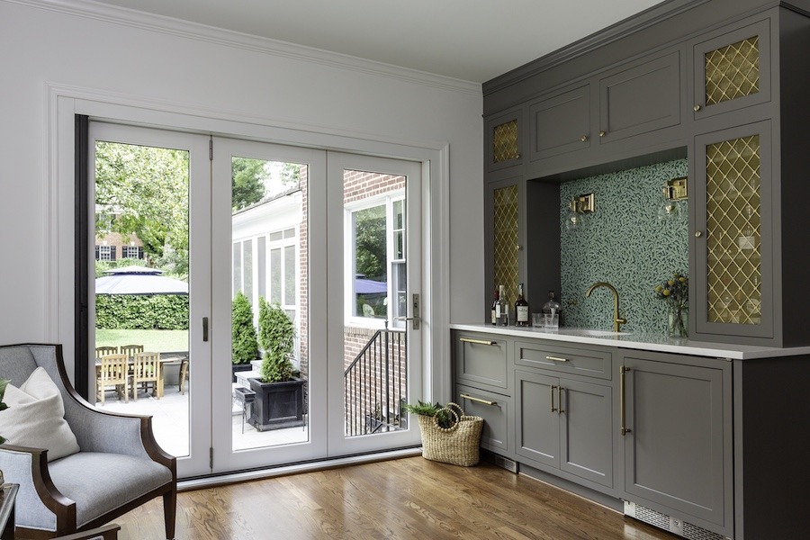 Large french doors bring light into living space with a large grey cabinet bar that features a green backsplash and gold accents.