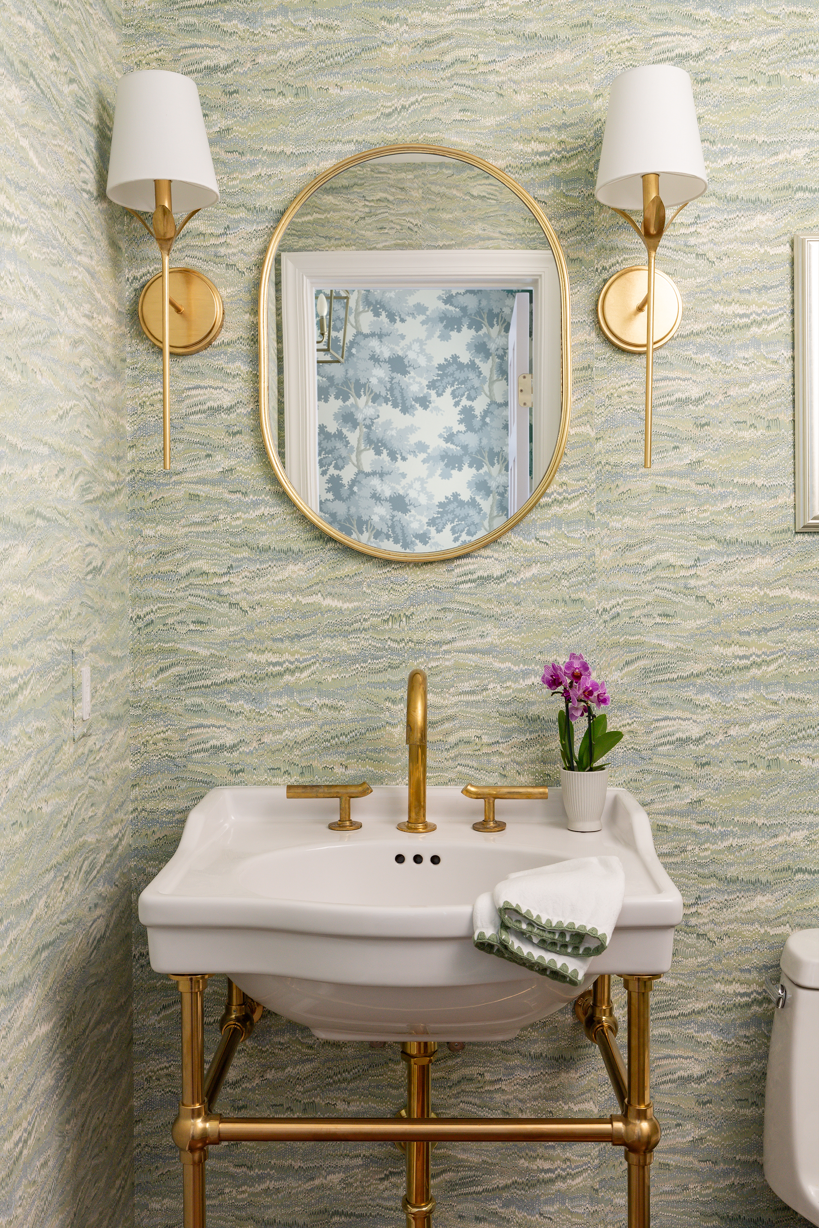 A mirror hanging above a sink in a bathroom, reflecting the interior design elements surrounding it, including sconces, a pedestal sink with bronze legs table and a flower decoration.