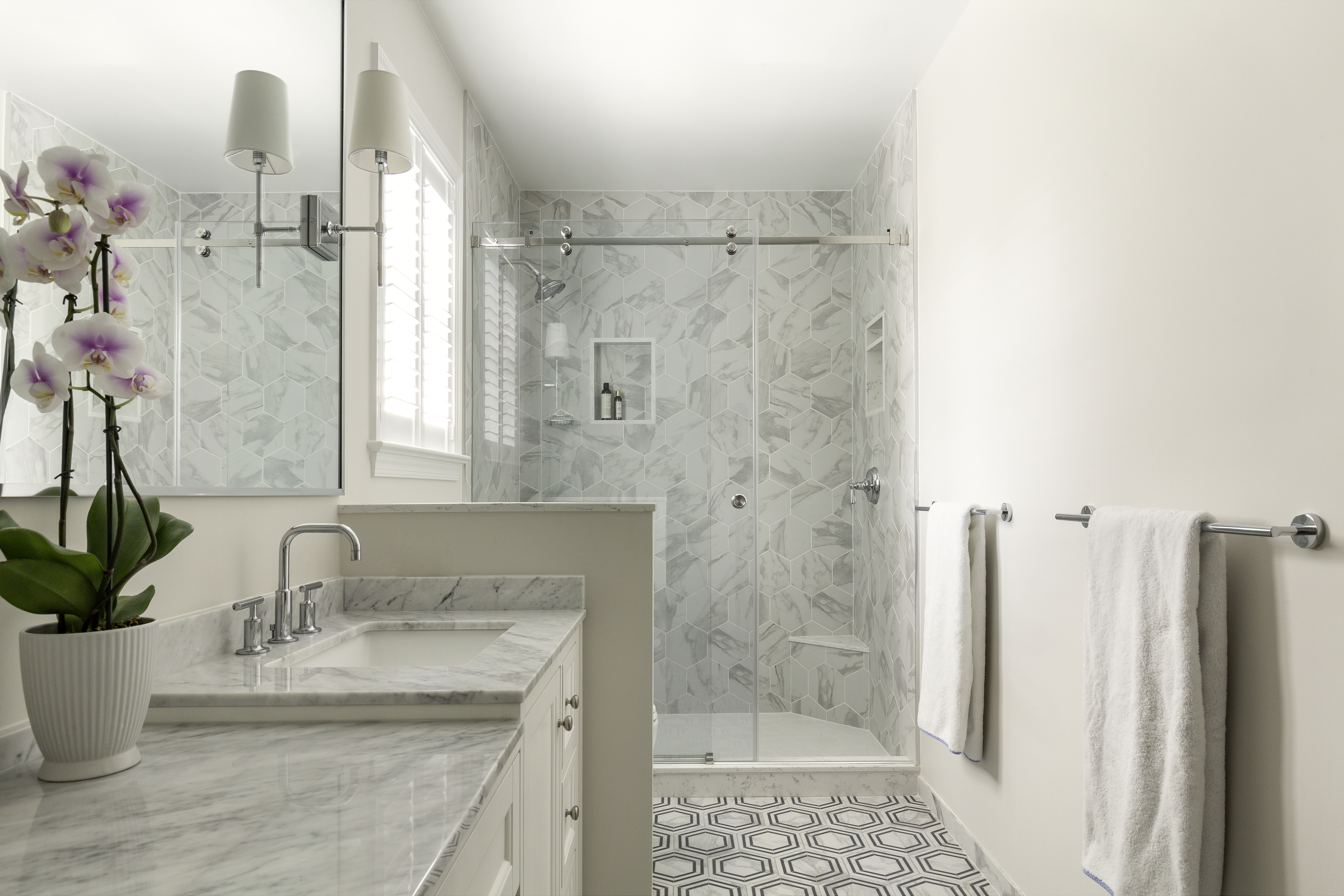 A bright bathroom featuring a shower and sink, with tiled walls and a countertop. A mirror is mounted above the sink, and a vase with flowers adds a touch of decor. The space is designed for comfort and functionality, with modern plumbing fixtures.