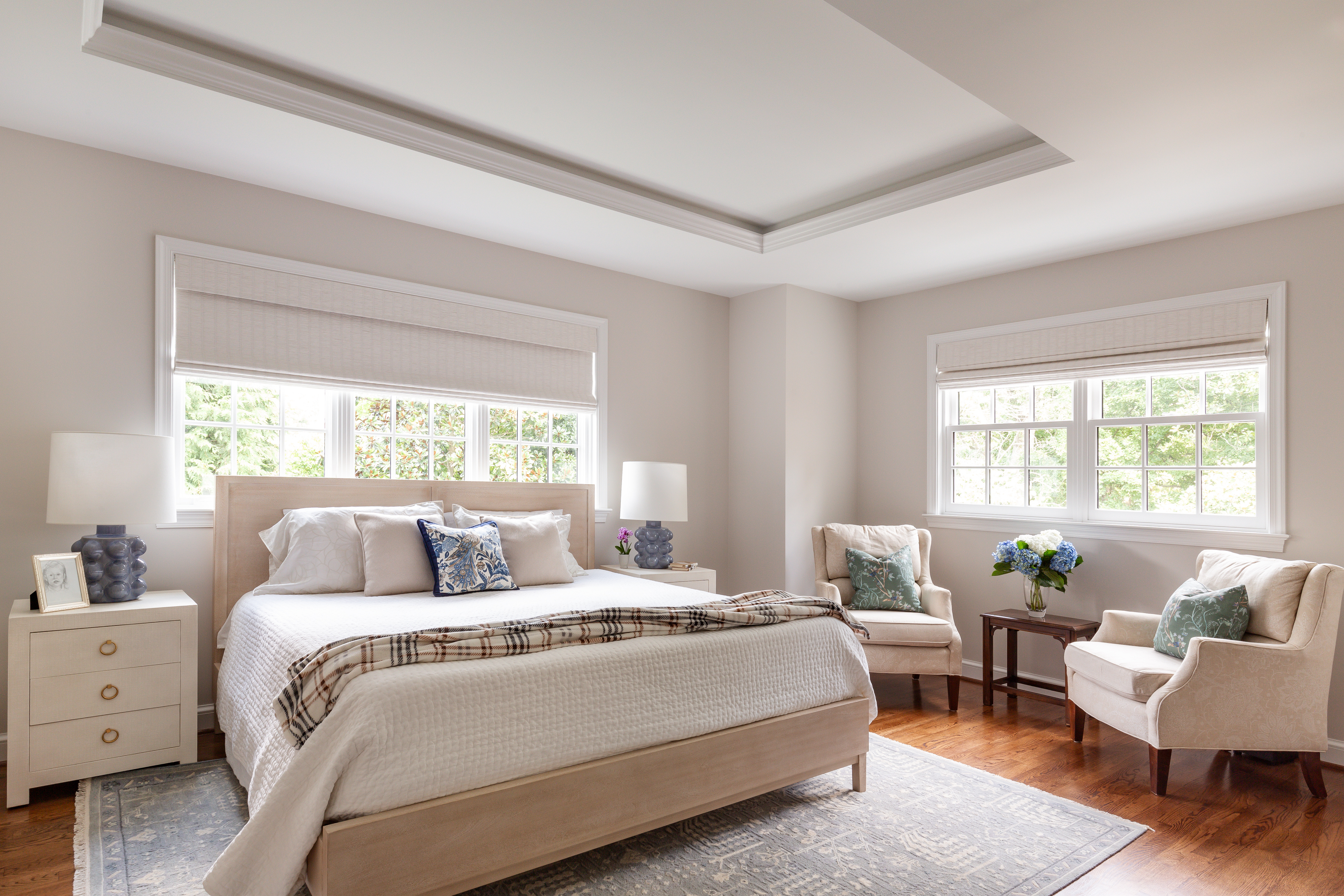 A stylish bedroom featuring large windows and a tray ceiling. There is a bed with decorative pillows, paired with chairs and a wooden nightstand. The room has hardwood flooring and a window with treatments, creating an inviting atmosphere.