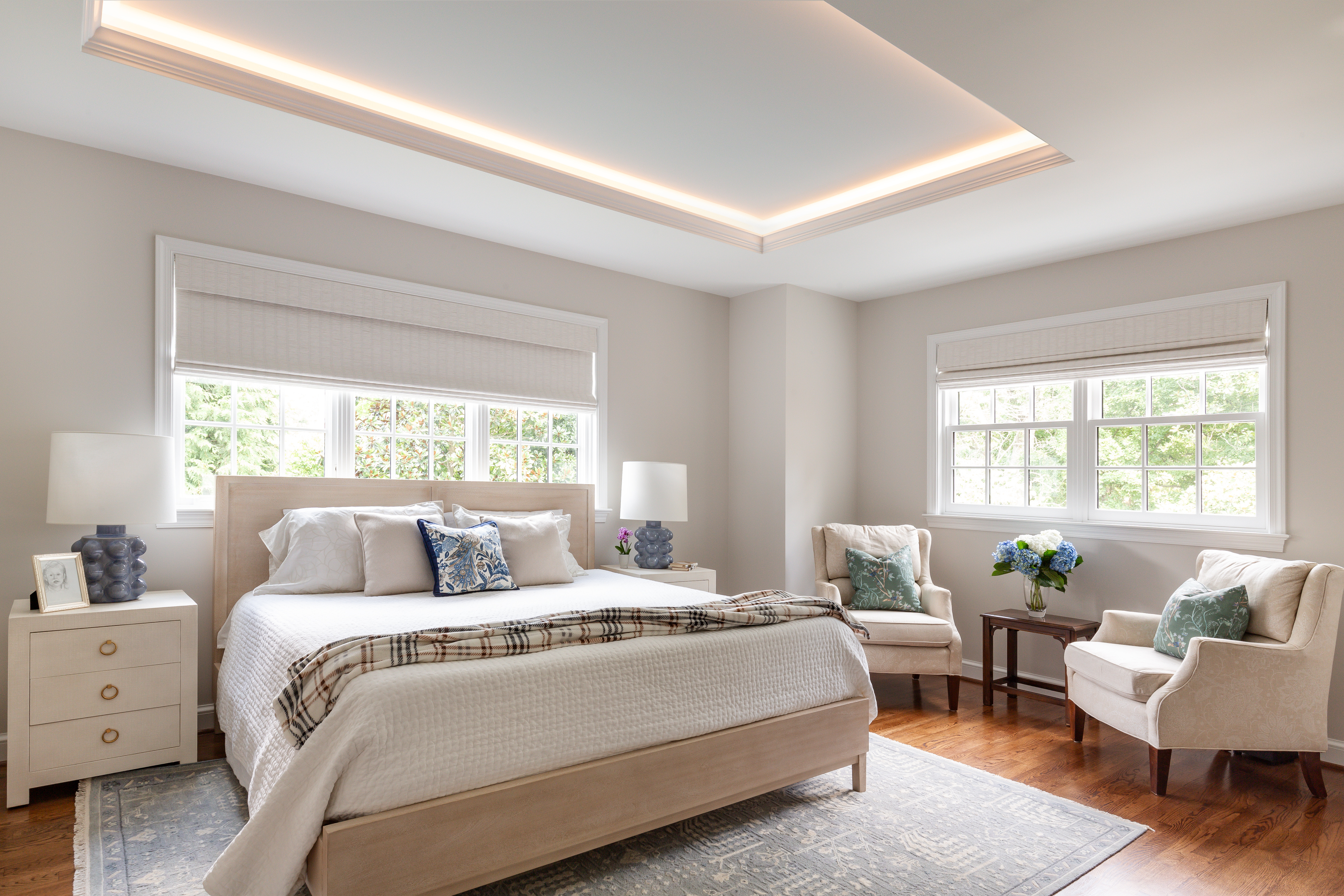 A cozy bedroom featuring a bed with pillows and a duvet cover, complemented by chairs. The room includes a nightstand and window treatments, with decorative cushions on the bed. The overall design showcases indoor interior elements such as molding, flooring, and ceiling details.