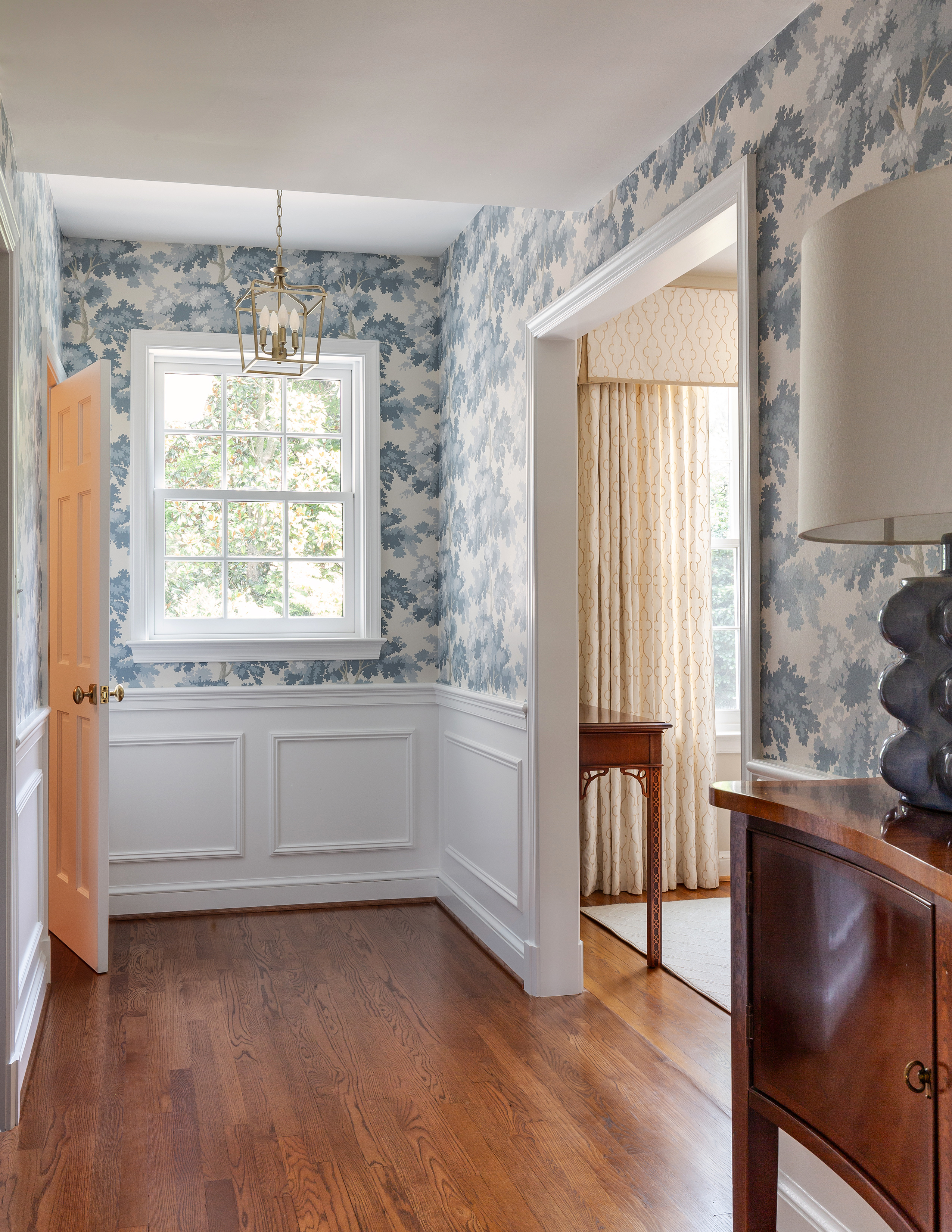 A well-lit entryway features a wood floor, white wainscotting and a large window, showcasing the interior design with wooden furniture and cabinetry. The ceiling is adorned, and a cozy window seat adds charm to the space.