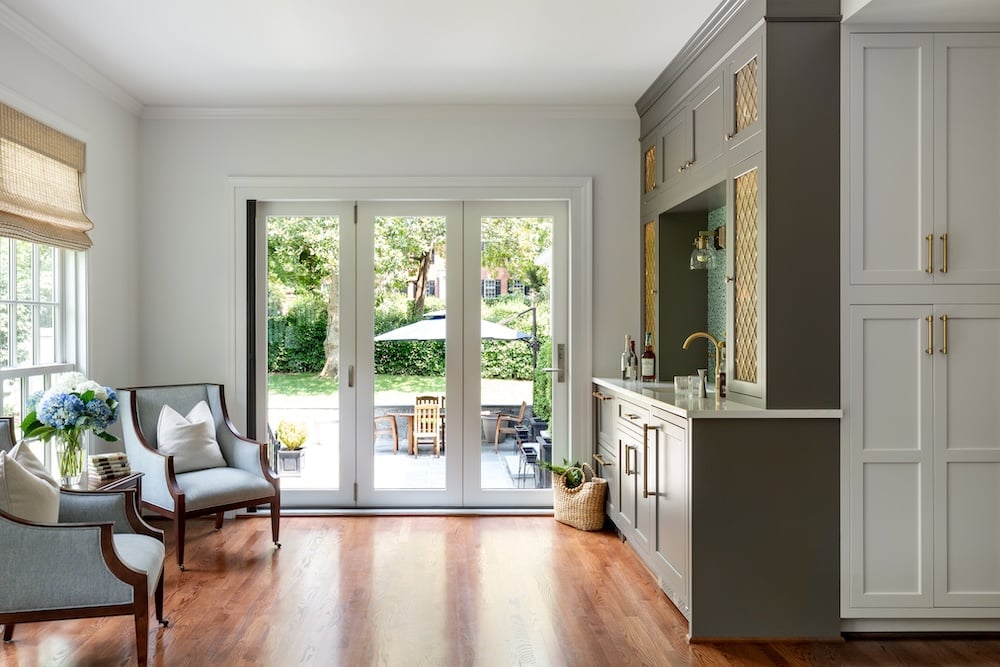 Bright, modern kitchen with hardwood floors, white cabinets, and gray accents from a recent Bethesda remodel. Glass doors open to a patio with outdoor seating. Two armchairs and a basket of flowers sit beside the windows.