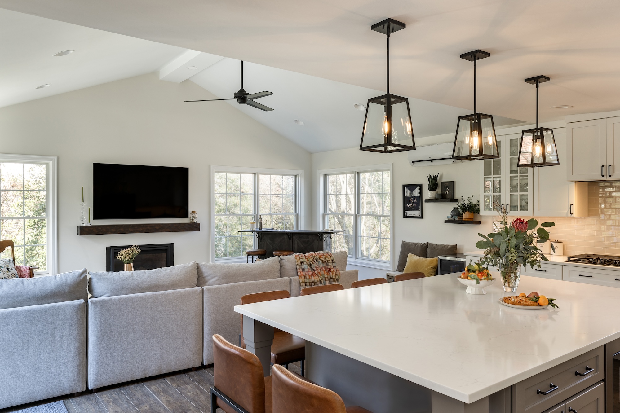 Modern open-concept living room and kitchen in Northwest DC, featuring a large island, pendant lights, gray sectional sofa, wall-mounted TV above a fireplace, and large windows—all crafted by an award-winning remodeler.