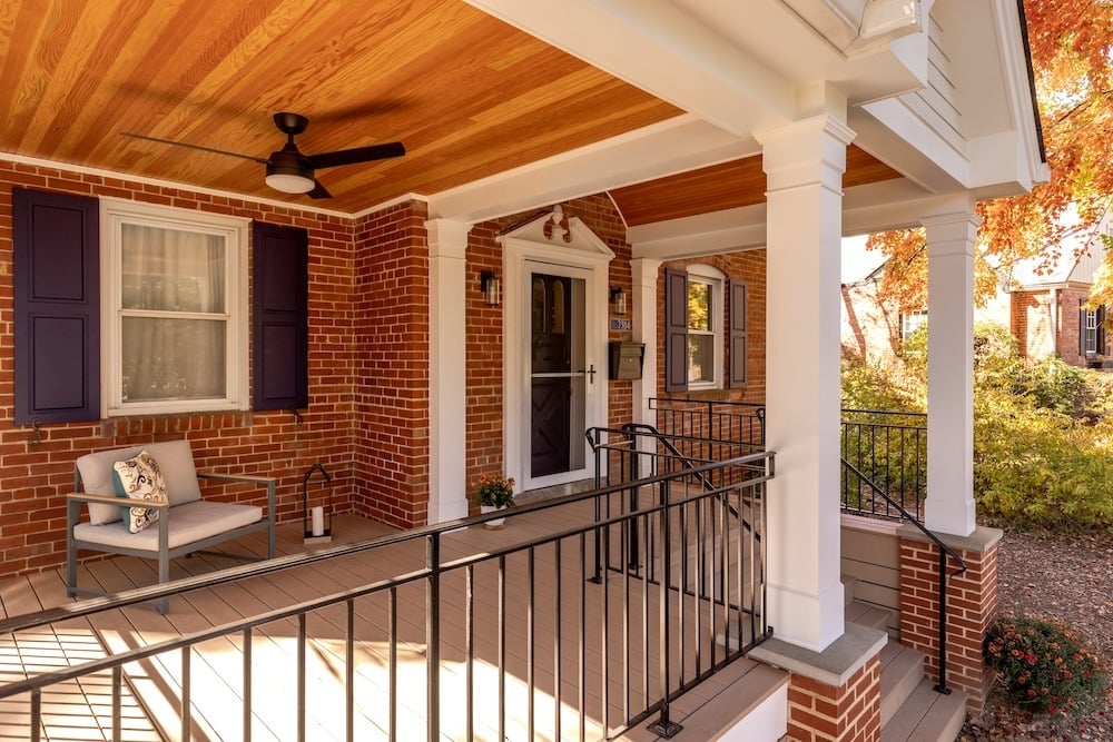 A cozy front porch with a wooden ceiling. It features a brick exterior, white pillars, and black railings. A cushioned chair with a pillow and a small plant sit near the door. Autumn foliage is visible in the background.