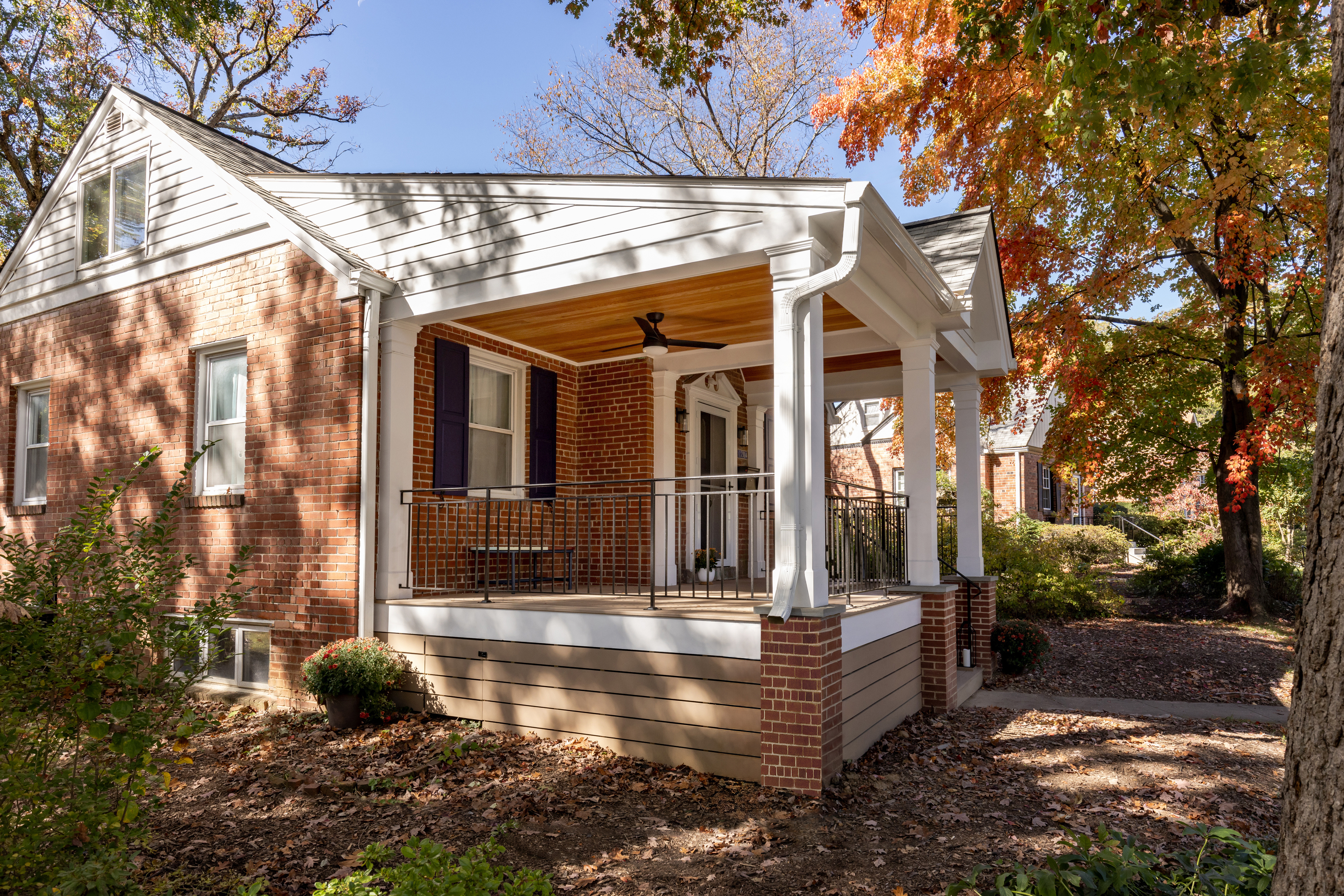 Trescott_After_FrA house with a large front porch featuring a vaulted ceiling, white pillars and black metal railing, is surrounded by trees and plants, featuring windows and a front door, with siding that suggests it's in a historic style. The ground is covered with fall foliage, adding to the outdoor ambiance.ontPorch1